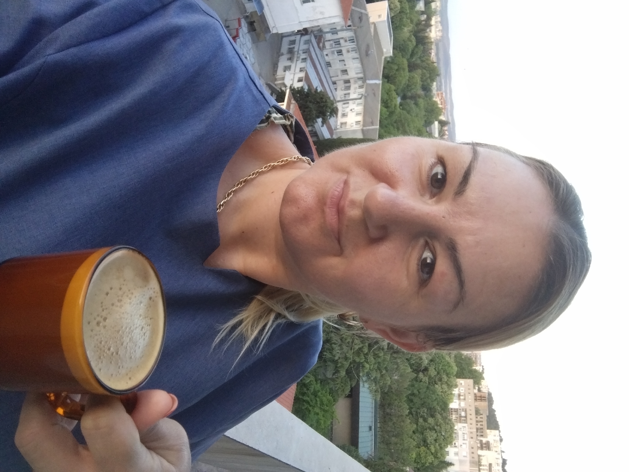 A woman is holding a mug of coffee with foam on top, outdoors, with a city view in the background. She is wearing a blue top and a gold chain. The background shows buildings and trees, typical of a city landscape. Based on the general architectural style and topography, the image possibly captures a scene in Stara Zagora, Bulgaria. More specific location cannot be determined.