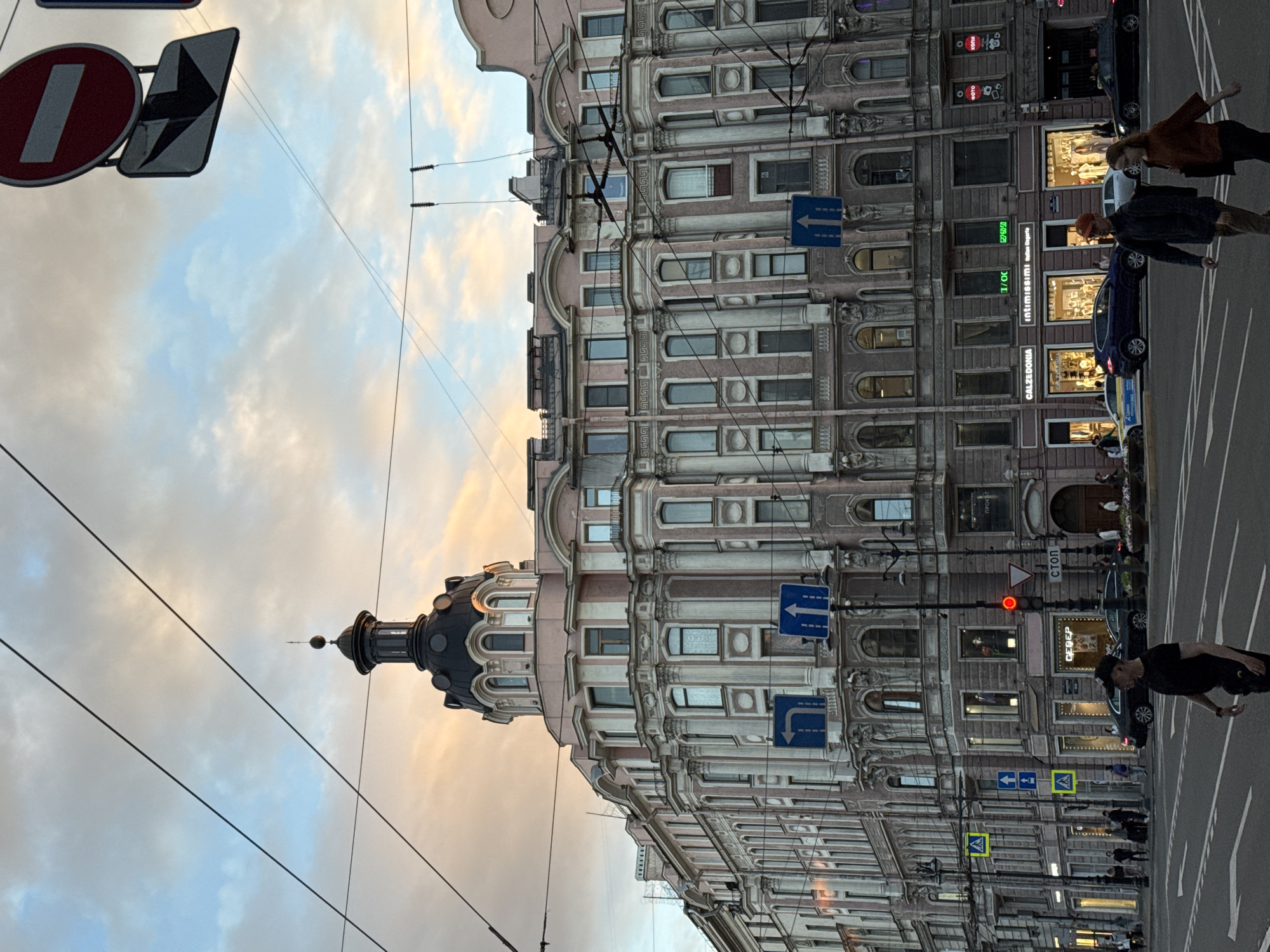 The image depicts a street scene in Saint Petersburg, Russia, captured during what appears to be sunset or twilight. Dominating the frame is a large, ornate building with classical architectural elements, including columns, intricate carvings, and a prominent dome. The building is painted in shades of pink and white, with numerous windows. Overhead, a complex network of electrical wires crisscrosses the sky.
Traffic signs are visible, including a "no entry" sign and directional arrows. A traffic light shows red. Several cars are on the street, some parked and others in motion. Pedestrians are walking across the street at a crosswalk. One pedestrian in the foreground is wearing a black t-shirt and a black baseball cap. Another pair of pedestrians, a man and a woman, are walking together further down the street. Shops with illuminated storefronts are visible at street level, with signage indicating brands like "CALZEDONIA" and "intimissimi." The sky displays a mix of clouds and the warm hues of the setting sun. The overall impression is of a busy urban environment with a blend of historic architecture and modern life.