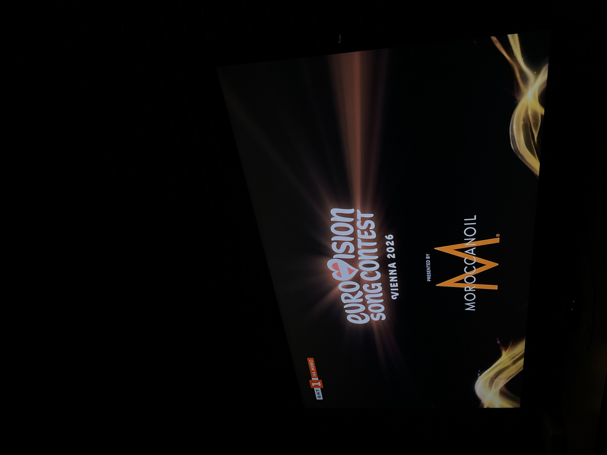The image shows a screen displaying the Eurovision Song Contest Vienna 2026 logo. The presenter is Moroccanoil. A television channel logo "БНТ 1 НА ЖИВО" is visible in the bottom left corner. The location is unknown.