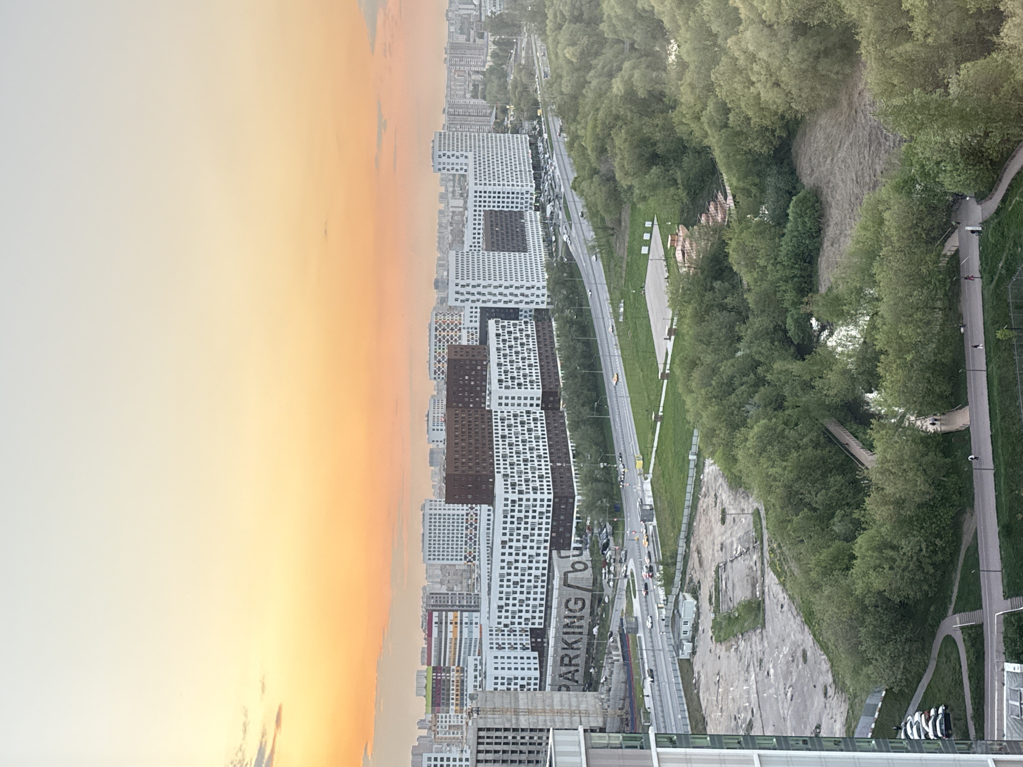 Here is a detailed description of the image:
The image shows a cityscape at sunset, with buildings, trees, and a road visible. The sky is a mix of yellow and orange, indicating sunset. On the left are multiple high-rise buildings with a mix of white and dark-colored facades, some still under construction. The signs near the buildings indicate "Parking". A highway runs along the length of the buildings, with moving cars. A green area with trees lines the right side of the road, suggesting a park or natural area. A walking path runs parallel to the road, and a walkway leads into the wooded area.