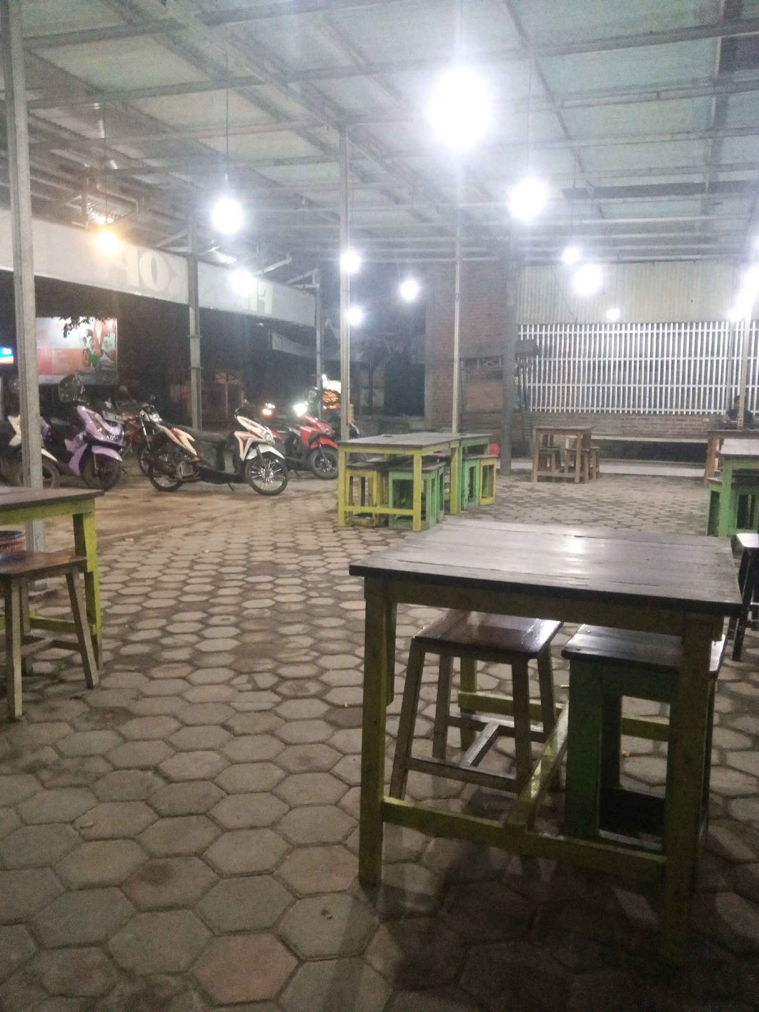 The image shows an outdoor seating area at night, likely a cafe or restaurant, in Gayaman, Indonesia. The area is covered by a metal roof supported by metal poles, with fluorescent lights illuminating the space. The ground is paved with hexagonal interlocking concrete tiles.
In the foreground, there are several wooden tables and chairs. One large wooden table with dark wooden stools is prominently featured, with a smaller green-legged table and stool to its right. In the midground, there are more green-legged tables and stools. Further back, a white metal fence encloses an area with wooden tables and chairs.
To the left of the seating area, several motorcycles are parked on the paved ground. These include a purple scooter, a white and black scooter, and a red scooter. Behind the motorcycles, a building with a sign that appears to say "KOFE" is visible. The overall atmosphere is dimly lit and suggests a casual, open-air dining or gathering space.