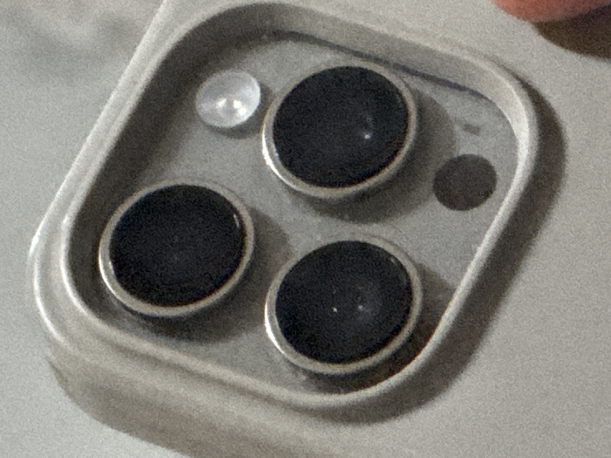 The image shows the camera array of a modern smartphone, likely an iPhone, with three lenses and an LED flash contained within a raised, rectangular housing on the back of the phone. The lenses are circular and black, with metallic rims. The LED flash is smaller and silvery. There is also a small dark circle within the camera array housing, likely a microphone. The phone itself appears to be light grey or silver in color. The image is close-up and does not reveal any identifiable location, including Plovdiv, Bulgaria. There are no people visible.
