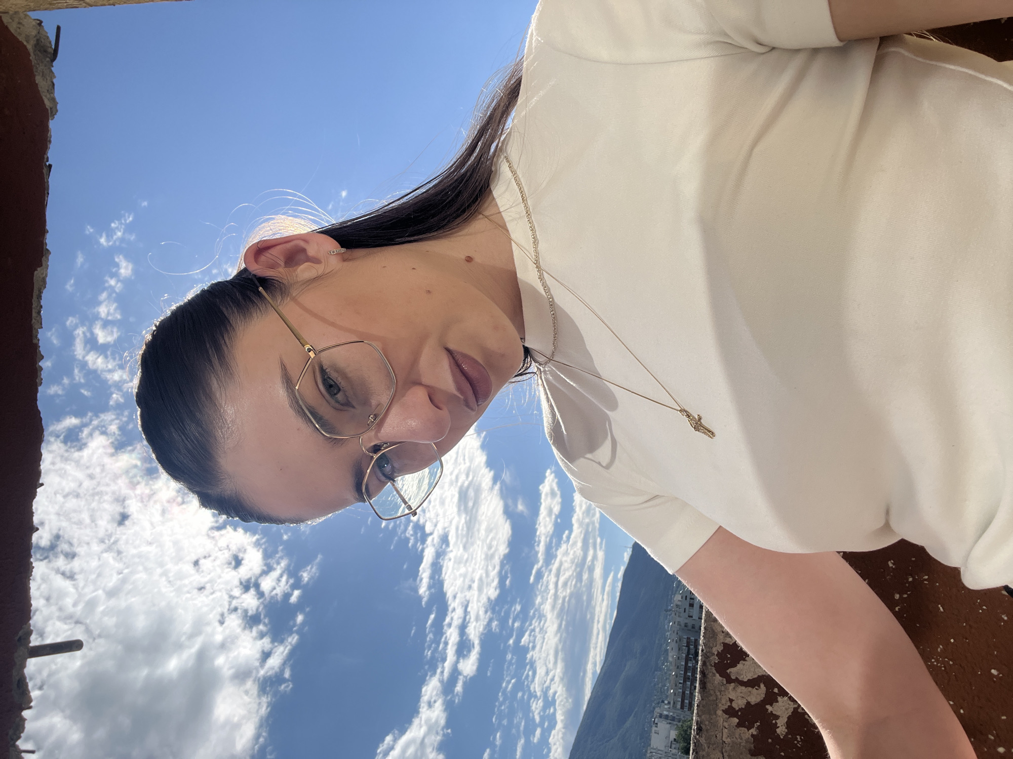 A low-angle, close-up, selfie-style shot shows a young woman with dark hair pulled back and wearing gold-rimmed glasses and a white t-shirt with a gold necklace featuring a cross pendant. She is outdoors with a bright blue sky and fluffy white clouds in the background, with mountains and a cityscape visible in the distance. The lighting suggests it is daytime and sunny.