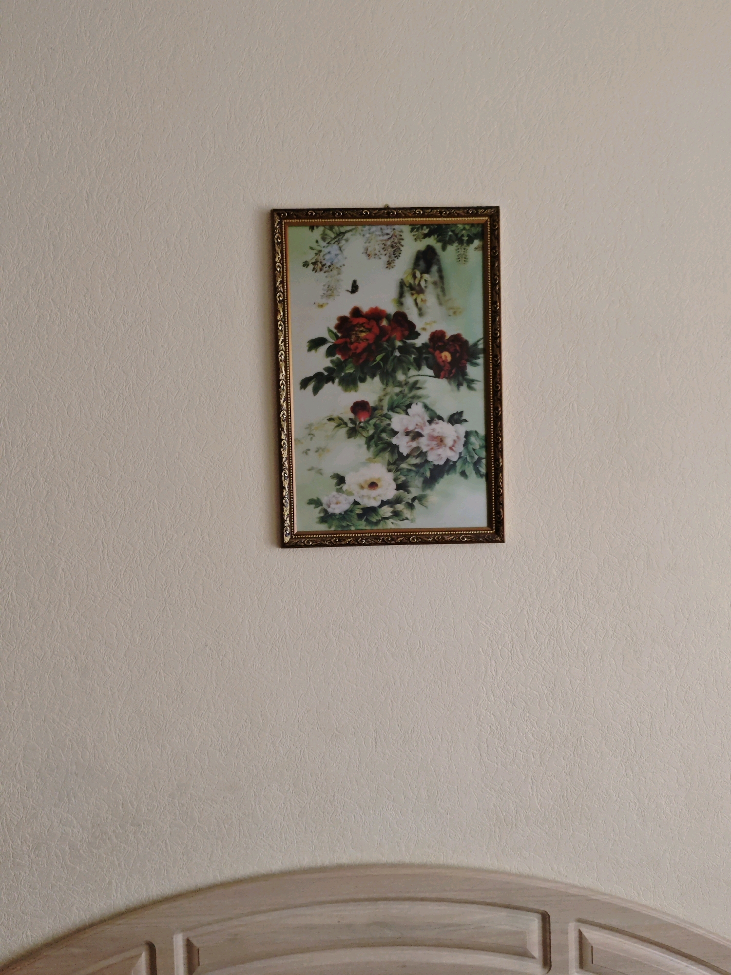 An ornate, framed painting of peonies and butterflies hangs on a textured, cream-colored wall in a room. The painting is rectangular and features a gold-colored frame with intricate detailing. The peonies are depicted in shades of deep red, pink, and white, with delicate butterflies flitting around them. Below the painting, the curved, light-colored wood of what appears to be a headboard is visible. The overall scene suggests an indoor setting, possibly a bedroom or living area, with a focus on decorative elements. The image does not provide any information about its location in Yevpatoriya, Ukraine.