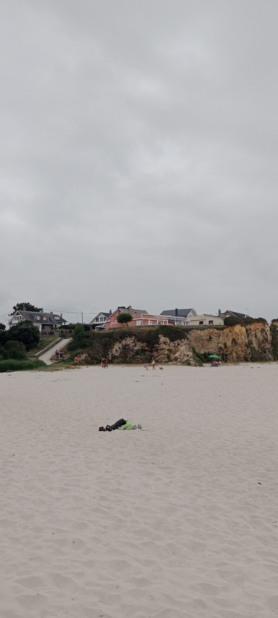 A wide-angle, vertical, medium-shot photograph captures a beach scene under a grey, overcast sky. In the foreground, a patch of light-colored sand is scattered with a few pieces of dark-colored exercise equipment, including what appears to be weight plates and a green bag, suggesting a recent workout or preparation for one.
The midground of the image features a sandy beach with several small figures engaged in various activities. Two small dogs are present, one appearing to chase the other. A few people are walking or standing on the sand, and further to the right, a person is seated under a green umbrella.
Dominating the background is a steep, rocky cliff face covered with vegetation. Perched atop the cliff are several houses with varying architectural styles and colors, including a prominent pink house with white trim. The houses overlook the beach and the sea beyond, though the water itself is not visible in this frame. A pathway with steps leads up the cliff from the beach, providing access to the houses. The overall impression is a slightly melancholic, cool, and breezy day at the beach with a residential area situated on the cliffs above. The scene is likely located in the city of A Rochela, Spain, known for its coastal scenery.
