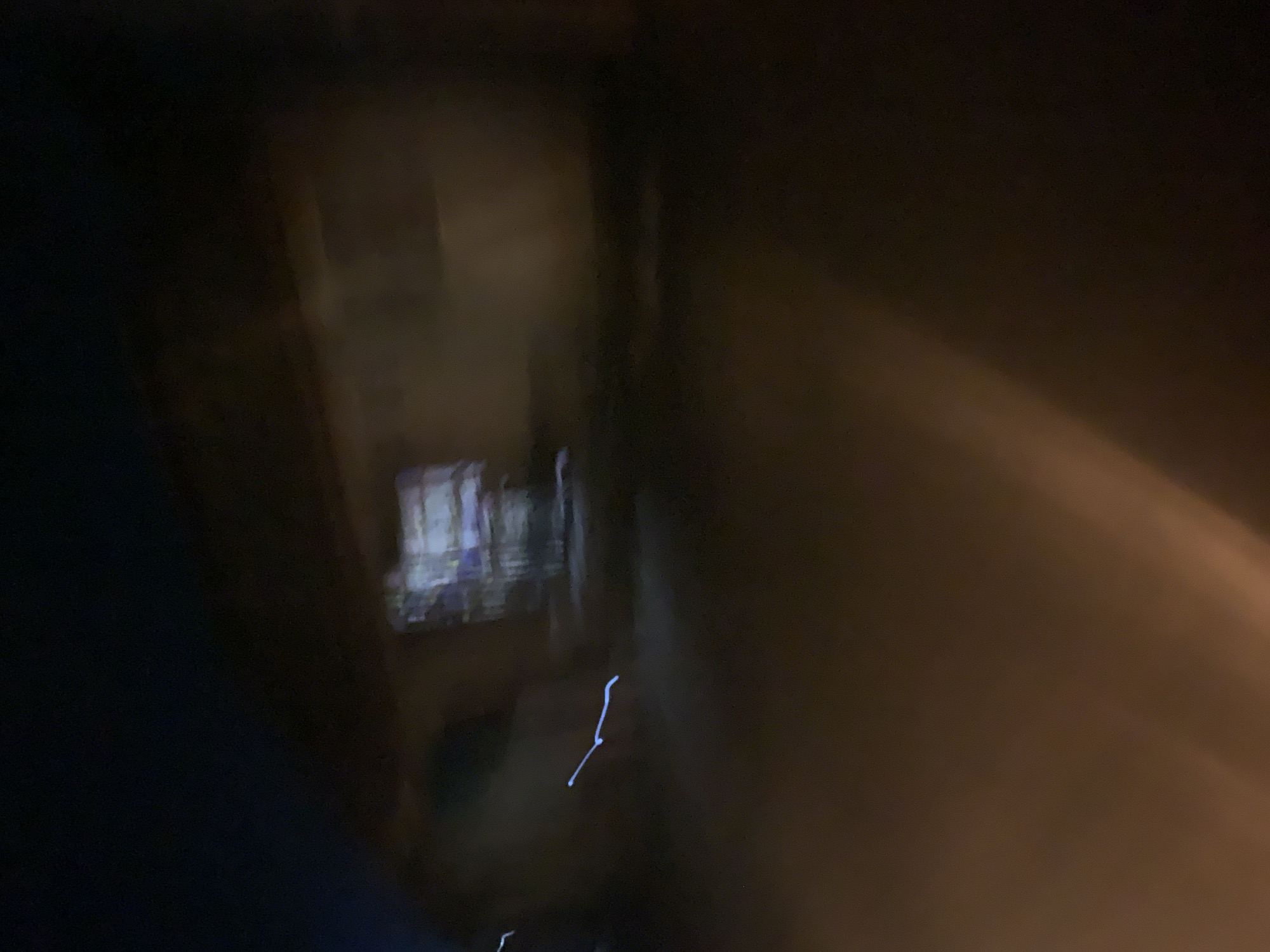 The image is a blurry, dark photograph of a building at night. The building appears to have illuminated windows. There is a streak of light in the foreground that may be from a vehicle or a light source. The ground is dark and appears to be wet. The image quality is poor, with significant motion blur.