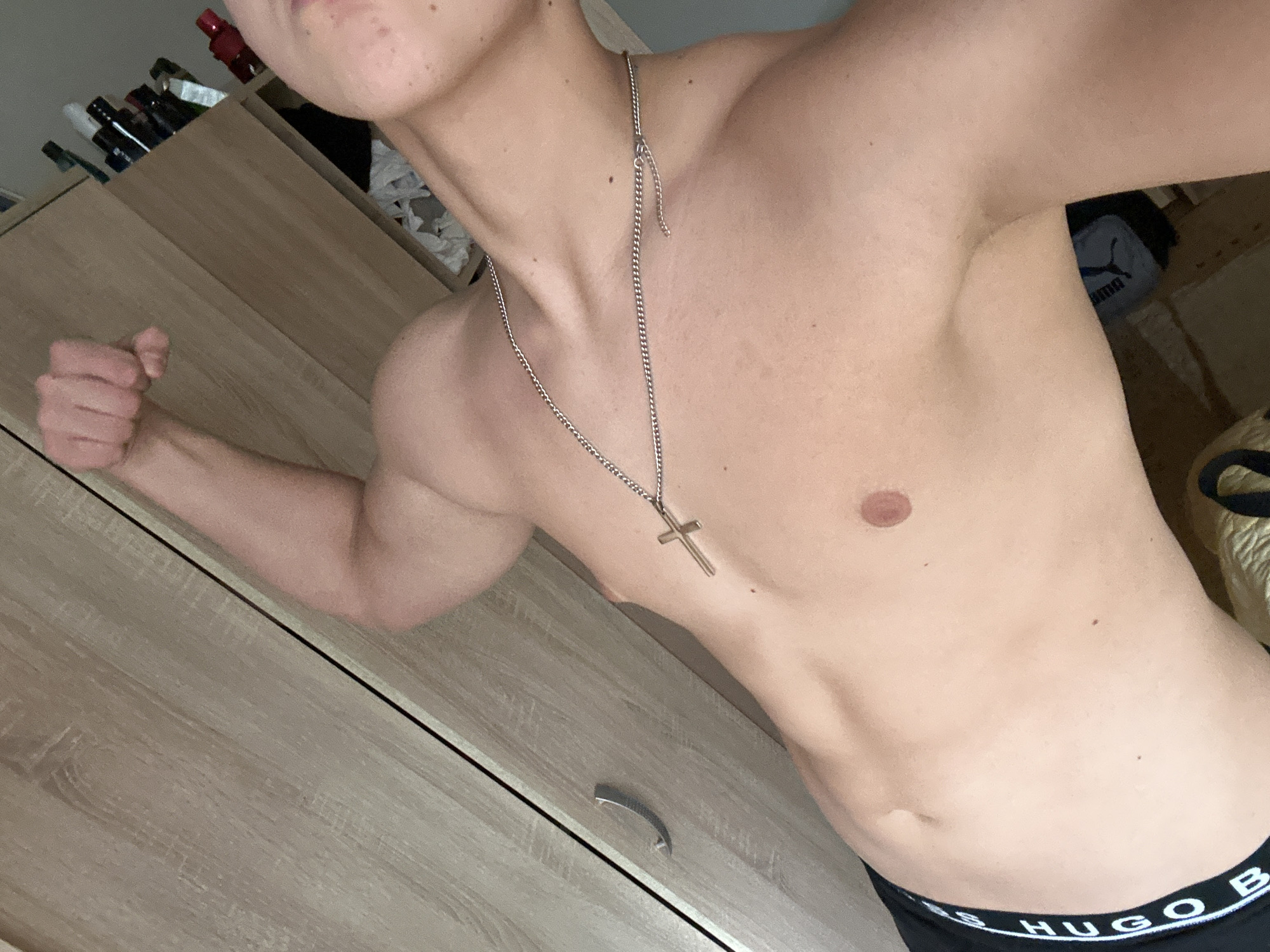 A shirtless individual wearing a silver cross necklace and black Hugo Boss underwear is flexing their left arm. The scene includes a wooden dresser with a silver handle and various objects on top, including what appear to be bottles. A PUMA bag is visible in the background. The location is not discernable from the image.