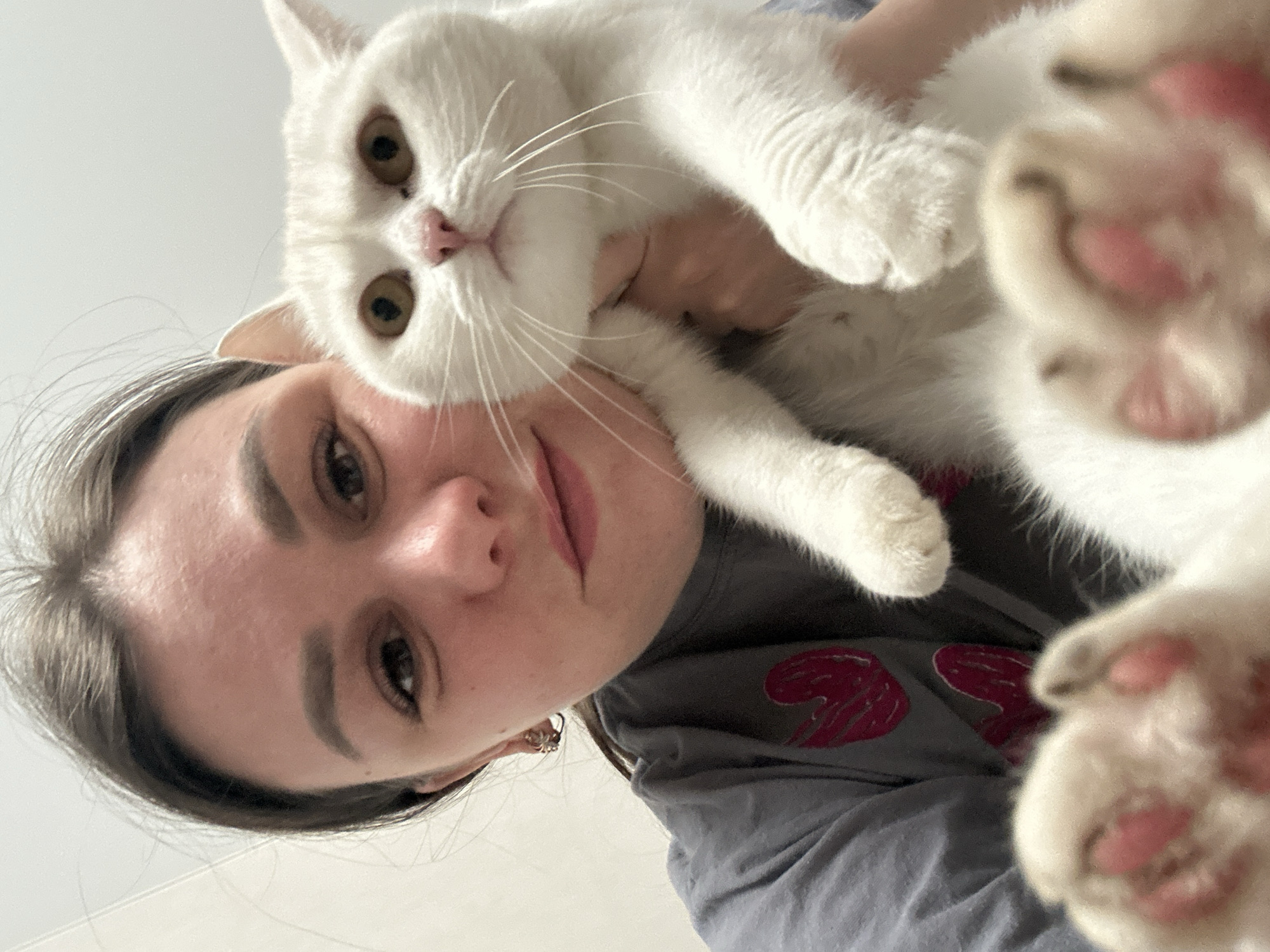 An intimate, low-angle selfie captures a young woman and a white cat in an indoor setting in Chaikovsky, Russia. The perspective looks upwards, with the woman's head and shoulders visible on the left and the cat positioned above her, closer to the camera.
The woman has dark hair and fair skin, with visible eyebrows and a gentle expression, looking directly at the camera with a slight smile. She wears a grey t-shirt adorned with a red, stylized winged design on the shoulder. A small, detailed earring is visible in her right ear.
The pure white cat dominates the upper right portion of the frame, looking intently at the viewer with large, round golden-amber eyes. Its pink nose and long white whiskers are prominent. One of its paws, with distinct pink paw pads, is blurry and very close to the camera, creating a sense of immediacy and playfulness. The cat's other front paw is also visible, resting near the woman's face. The cat appears to be held or resting comfortably on the woman, suggesting a close bond.
The background is a plain, light color, likely a ceiling or a simple wall, providing a clean backdrop for the subjects. The lighting is bright and even, indicative of daytime indoors. The image conveys a warm, affectionate, and playful moment between the woman and her feline companion. There is no visible text in the image.