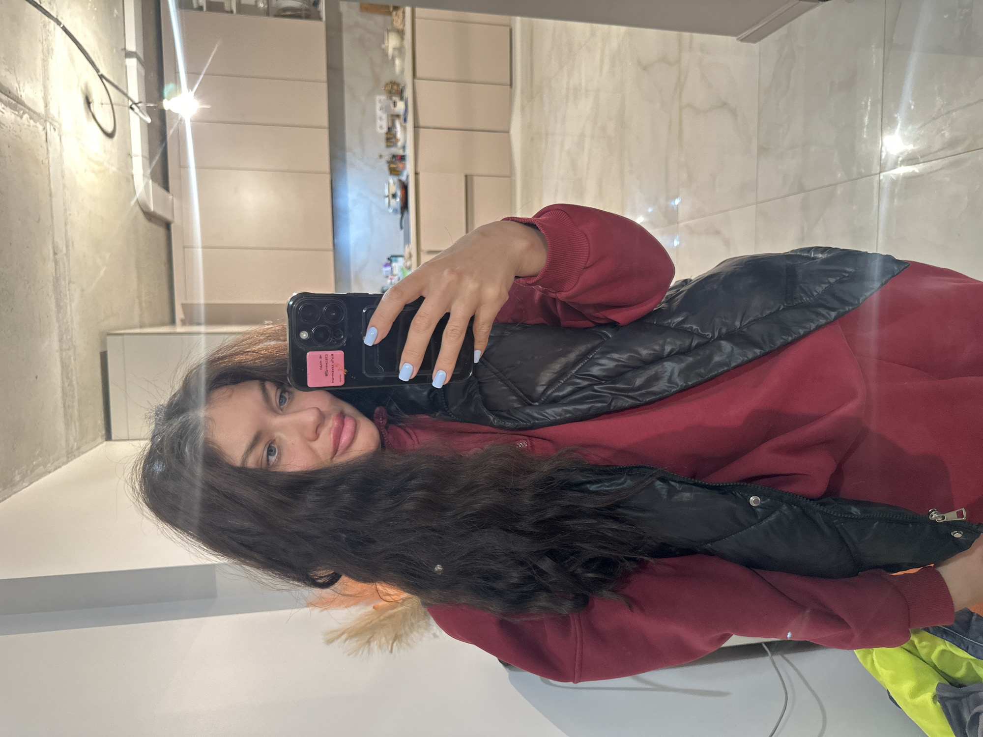 A woman with long, dark hair is taking a selfie in what appears to be an apartment or house interior. She is wearing a dark red sweatshirt and a black vest. She is holding a black smartphone with light blue nail polish on her fingers. The background shows a kitchen area with cabinets and what looks like marble-patterned wall or floor tiling. There is a light fixture hanging from the ceiling. The location is likely inside a building, but without additional information, it is impossible to confirm if it is in Khimki, Russia.