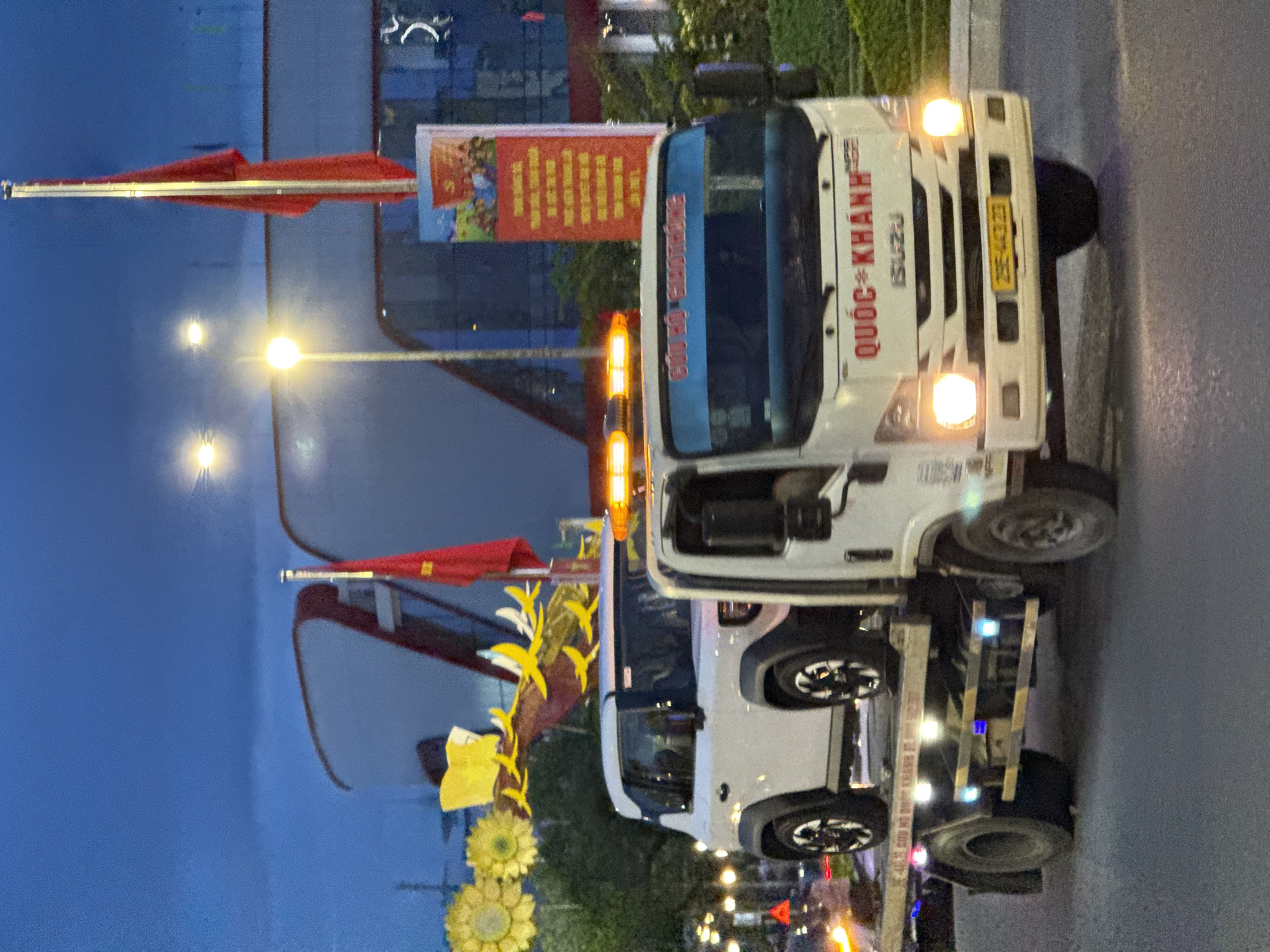 The image shows a tow truck with a white SUV on its flatbed, driving on a street in Nha Trang, Vietnam. The tow truck has orange flashing lights on its roof and a banner with Vietnamese text that reads "CỨU HỘ GIAO THÔNG QUỐC KHÁNH ISUZU" and a license plate "29E-443.23". The SUV appears to be a modern white model with black trim. In the background, there are large modern buildings, streetlights, and decorative yellow flowers. Two red flags are visible, one on a flagpole and another attached to a pole behind the SUV. The sky is dark blue, indicating it is either early morning or late evening. The scene suggests a normal street activity, possibly a vehicle breakdown or transport.