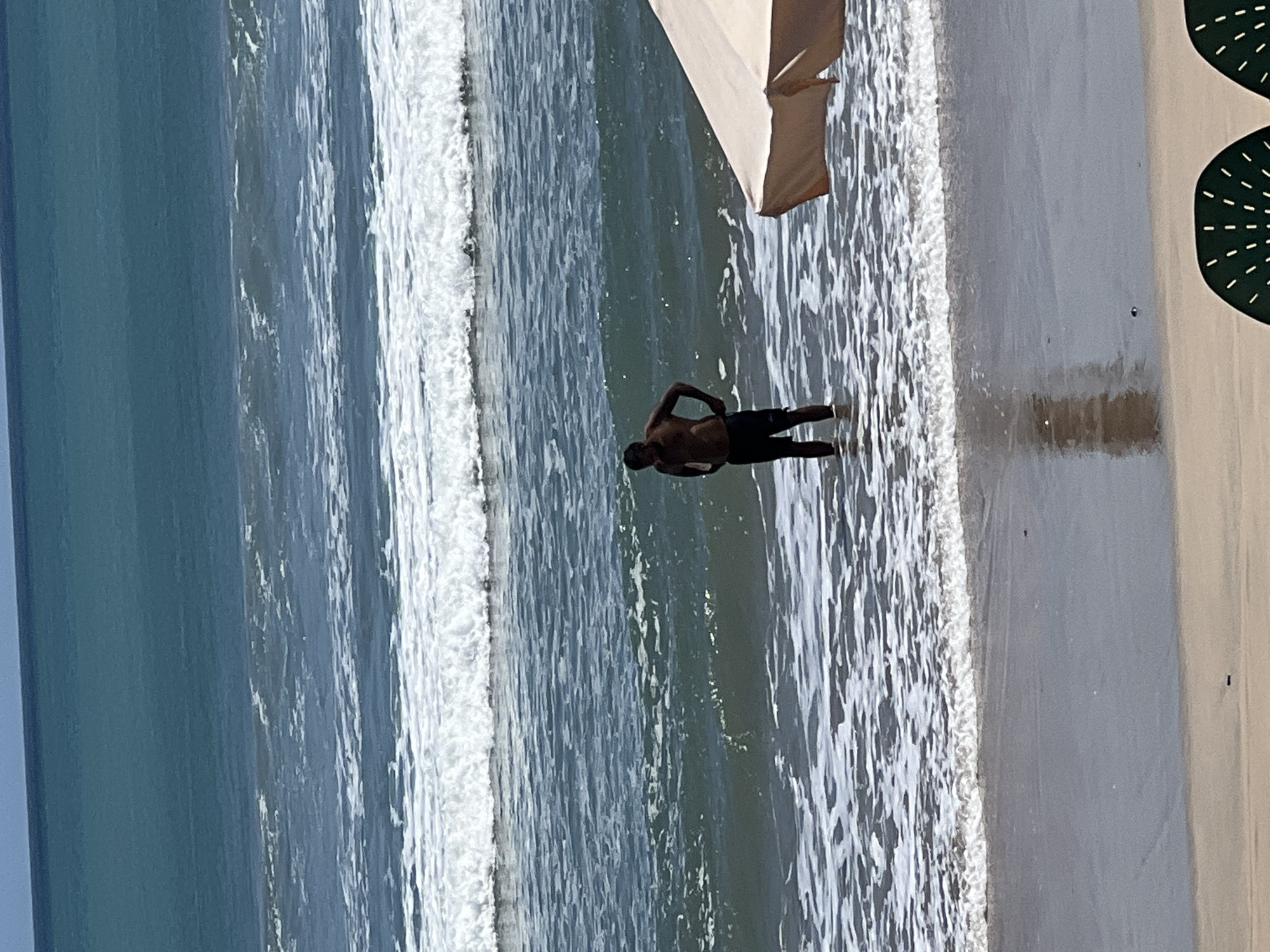 A man stands waist-deep in the ocean water near the shore, his hands on his hips. He is wearing black shorts. In the foreground, a portion of a beach umbrella is visible, as are two dark green beach chairs with white speckles. The ocean has several rows of waves, with white foam cresting on some. The sky is a clear blue. The scene is the beach in Kuta, Indonesia.