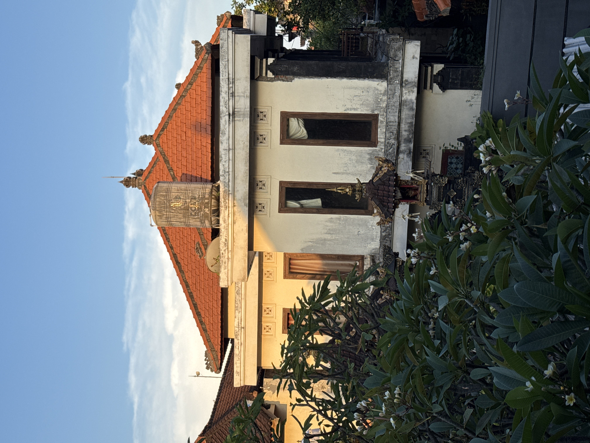 A sun-kissed, multi-story building featuring traditional Balinese architecture stands under a clear blue sky with wispy white clouds. The building's prominent red-tiled roof is adorned with ornate, terracotta finials along its ridge and peaks. A large, cylindrical metal water tank is visible on the roof, along with a small satellite dish. The facade of the building is painted a light cream or off-white, showing signs of weathering and age, particularly below the roofline and around the windows.
The building features several rectangular windows with dark frames, behind which curtains are visible, some light-colored and others darker. Between the windows, square decorative panels with intricate patterns are inset into the wall. To the right of a lower window, a smaller, highly ornate structure with a dark, tiered roof and golden accents suggests a traditional Balinese shrine or decorative element.
Lush, dark green foliage, likely from plumeria (frangipani) trees, occupies the right and lower foreground, partially obscuring parts of the building. Numerous small white plumeria flowers are scattered among the leaves. The lighting suggests the golden hour, possibly late afternoon, as warm sunlight illuminates parts of the building's facade, casting distinct shadows and highlighting textures. The architectural style, decorative elements, and plumeria trees strongly indicate a setting in Bali, consistent with Kuta, Indonesia. There are no people or discernible text visible in the image.