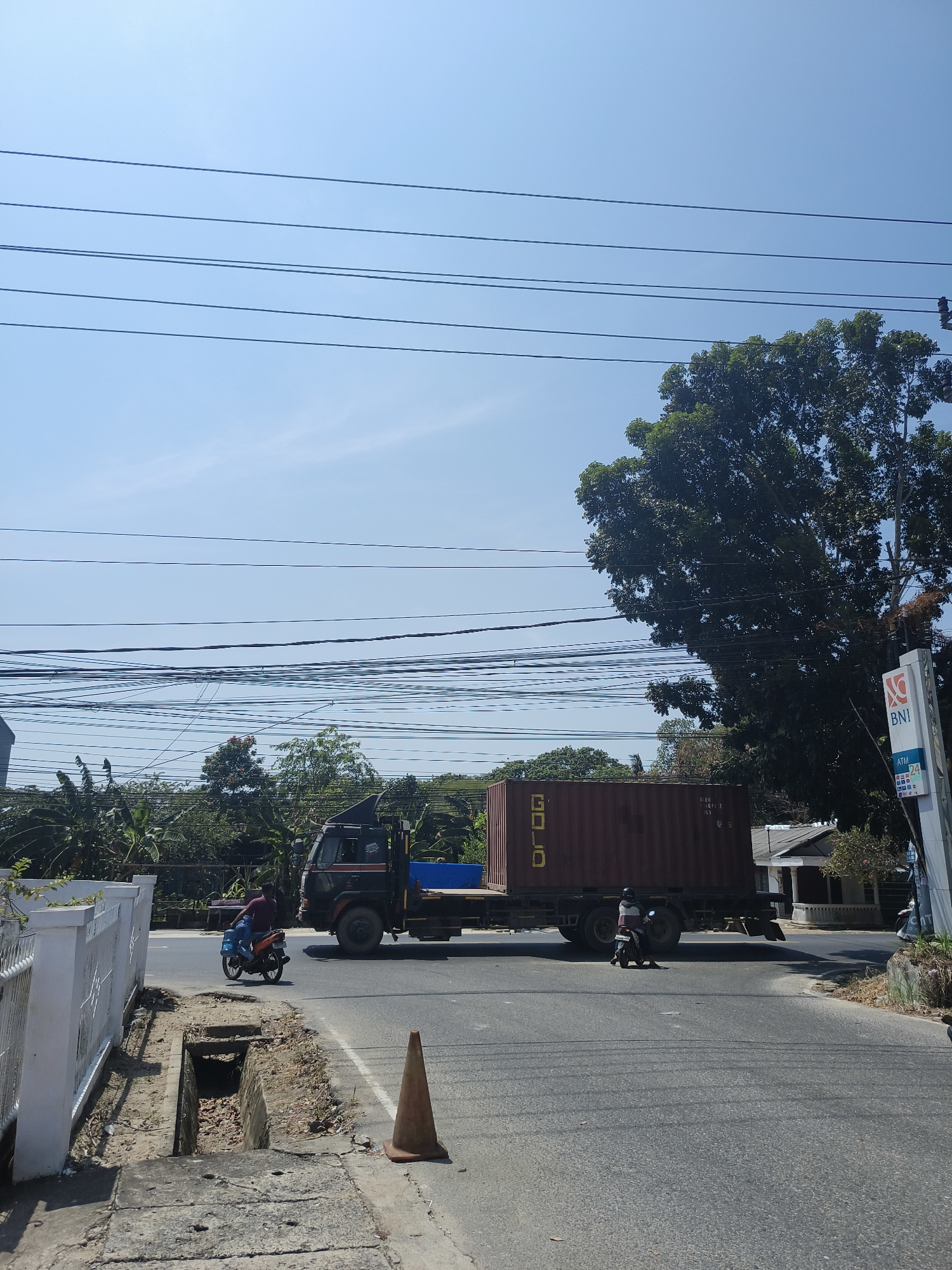The image depicts a street scene in Pangkalpinang, Indonesia, under a clear blue sky. In the foreground, a road runs diagonally from the lower left to the upper right. On the left side of the road, a white fence borders a drainage ditch filled with debris and dry leaves. An orange traffic cone is placed on the asphalt near the ditch.
A person on an orange motorcycle is traveling away from the camera on the left side of the road. Further down the road, a large, dark truck with a red shipping container is moving from left to right. The shipping container has the word "GOLD" written on its side in yellow lettering. Another person on a dark motorcycle is following the truck.
In the background, lush green trees and vegetation are visible. A tall, leafy tree stands prominently on the right side of the frame. To the right of the tree, a sign for "BNI" (Bank Negara Indonesia) with an "ATM 24" indication is mounted on a pole. Several utility poles with numerous power lines crisscross the sky. Buildings are partially visible behind the trees. The scene appears to be a typical day on a suburban or rural road in Pangkalpinang.