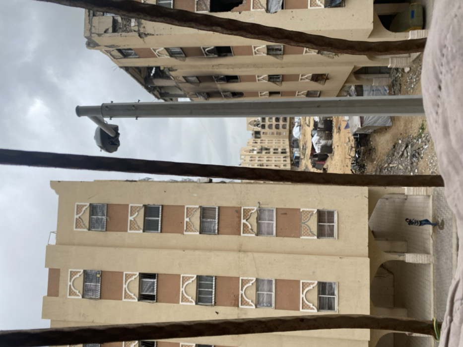 Damaged buildings stand in the background, marred by destruction. In the foreground, similar structures exhibit rows of windows adorned with decorative accents. A light pole extends into the frame, positioned between the buildings. Rubble and debris fill the open space between the buildings. A person is seen on the ground in the lower right corner. Weather looks overcast.