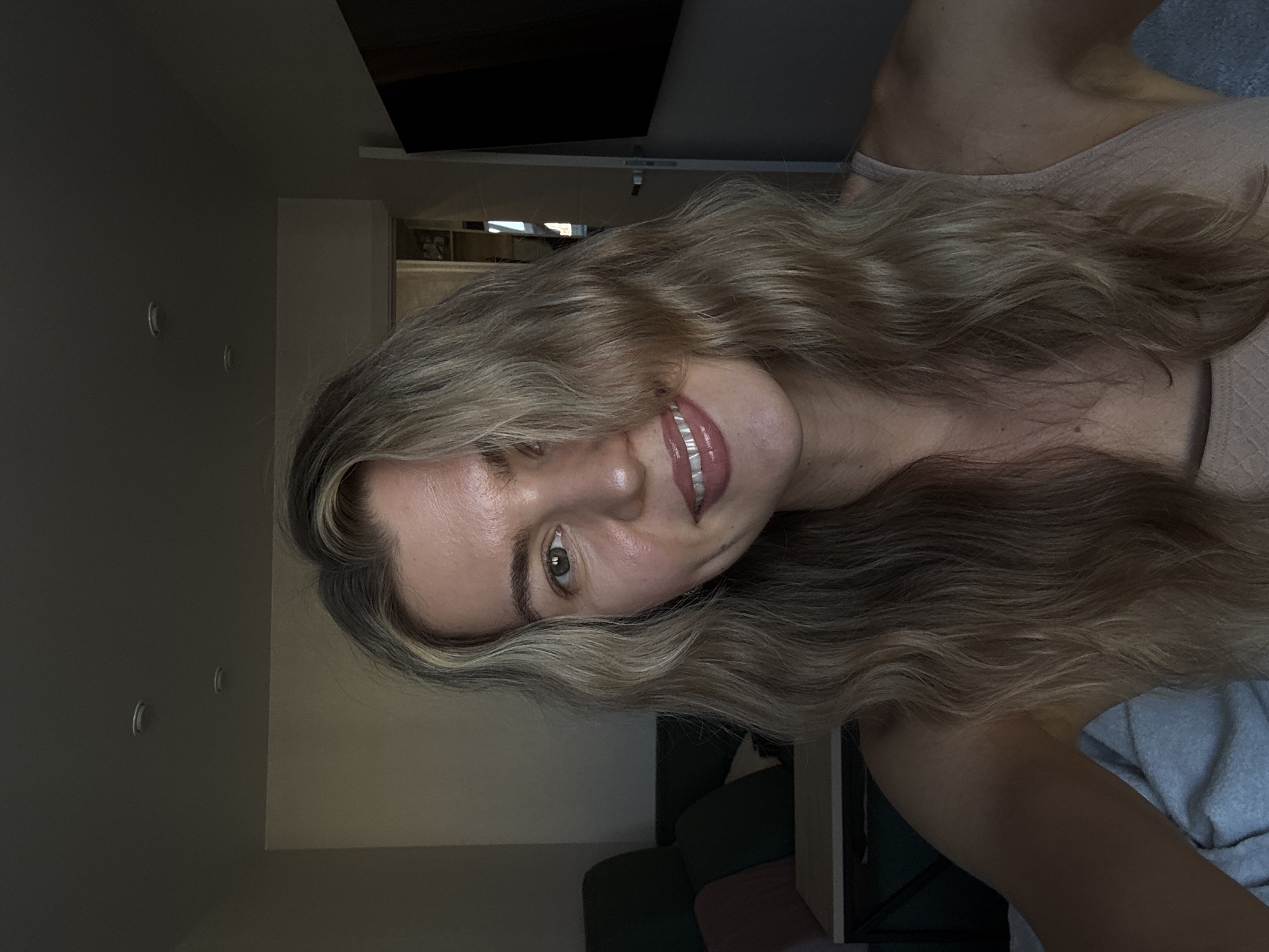 The image displays a headshot selfie of a young woman with long, wavy blonde hair, rotated 90 degrees clockwise. She is smiling softly, looking towards the camera with her head slightly tilted. Her hair, a mix of blonde and light brown tones, cascades over her left shoulder and partially frames her face. She has light-colored eyes, possibly green or hazel, and her lips are glossy. She is wearing a light pink or beige ribbed tank top or camisole.
The setting is indoors, appearing to be a room such as a living area or bedroom. To the left of the woman (when upright), a plain, light-colored wall is visible, featuring two small, circular, light-toned fixtures, which could be hooks or recessed lights. Above these fixtures, a dark, rectangular screen, likely a television, is mounted high on the wall. To the right, the background shows a hint of a dark green piece of furniture, possibly a sofa or chair, and a light wooden surface. A light blue blanket or fabric is also visible in the lower right corner. The lighting is soft and diffused, creating a relaxed ambiance. No text is visible in the image.