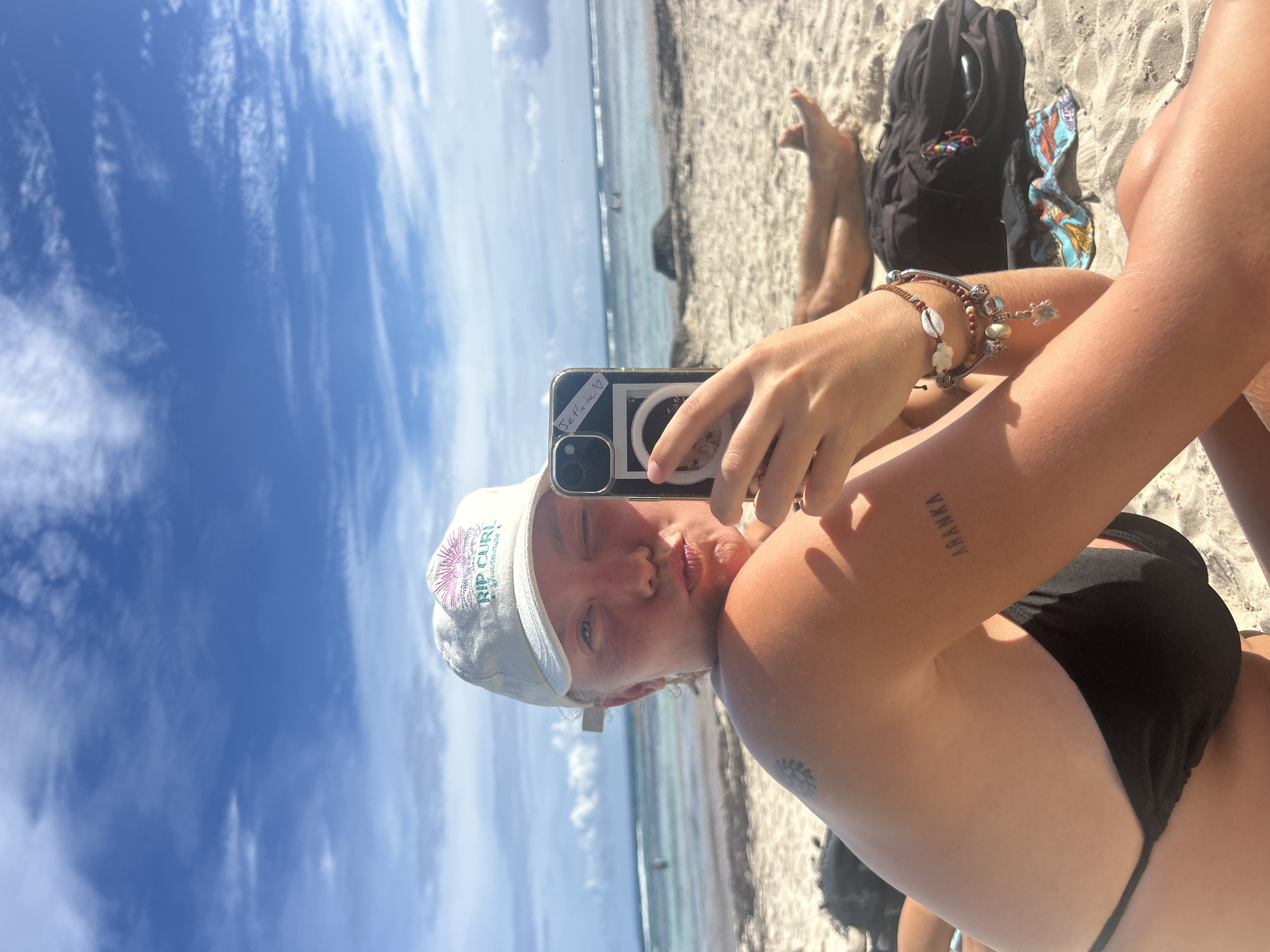 The image depicts a woman taking a selfie on a beach. She is wearing a Rip Curl cap and a black bikini top. She has multiple bracelets on her wrist and a tattoo on her arm that reads "Ayanka". She is holding a smartphone to take the picture. In the background, there is a sandy beach, the ocean, and a partly cloudy sky. A black backpack sits behind her. Given the prompt's indication of Sainte-Anne, France, the beach is likely located there.