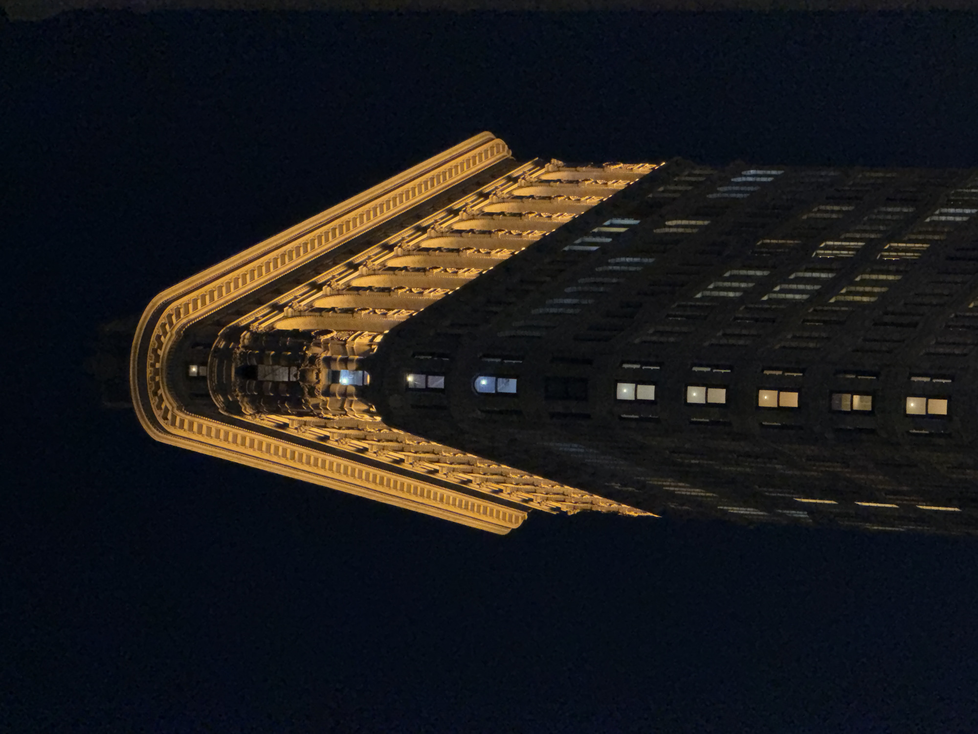 This is a low-angle, nighttime photograph of the Flatiron Building, a landmark skyscraper in Manhattan, New York City. The building's distinctive triangular shape is emphasized by the low vantage point and the dark night sky. The facade is illuminated by warm, yellow lights, highlighting its architectural details, including rows of windows and decorative elements along the upper floors. The building is silhouetted against the deep blue-black of the night sky, with only a few scattered lights visible in the distance. No people are visible in the frame. The focus is solely on the architecture of the building.
