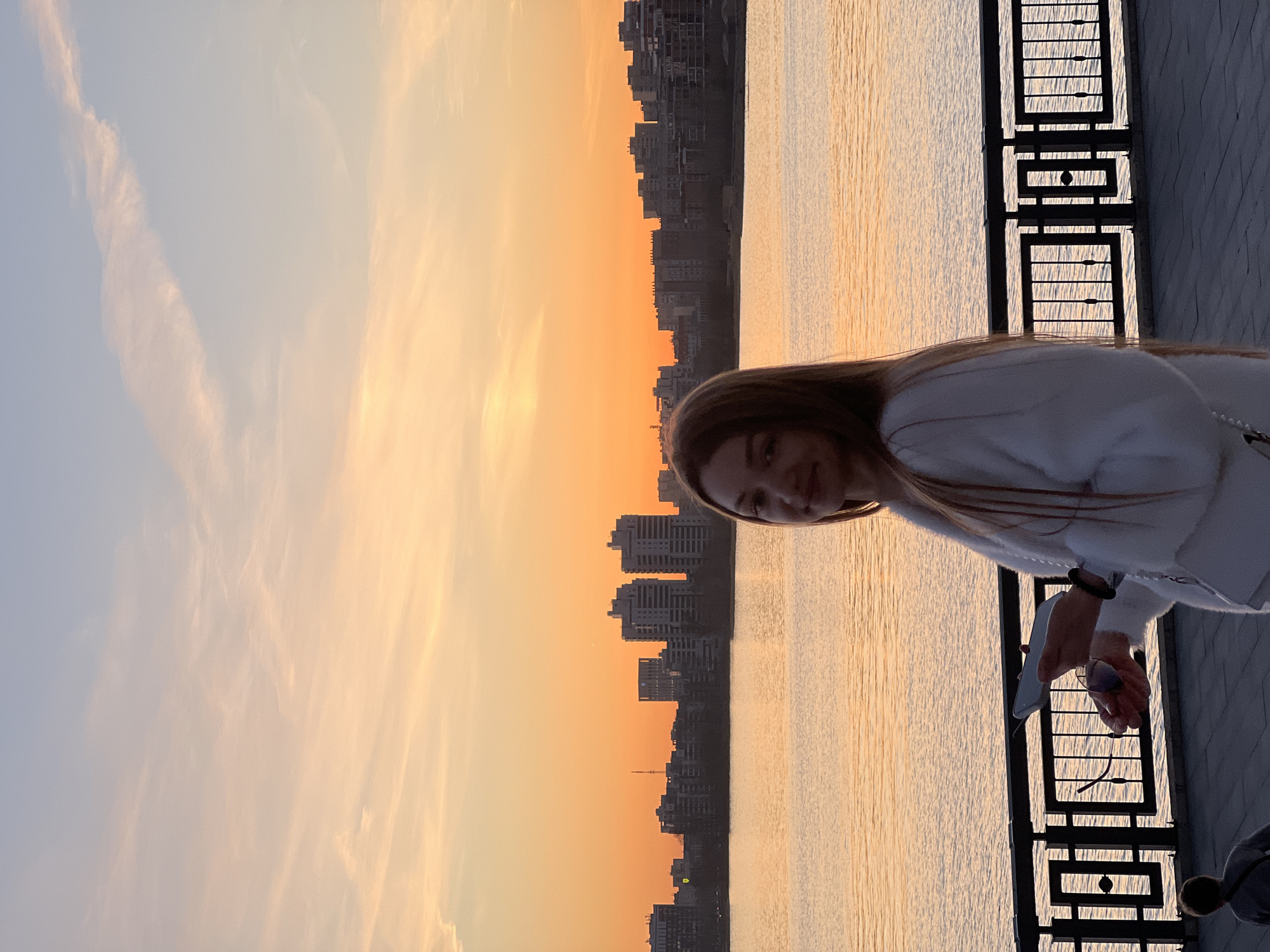 This image depicts a woman enjoying a picturesque sunset or sunrise over a city and body of water in Kazan, Russia. The photograph is oriented vertically, with the main subject, a young woman, positioned on the right side of the frame. She has long, light brown hair and is wearing a light-colored, possibly white, sweater. She is leaning against a dark, ornate metal railing, looking towards the camera with a slight smile. In her hands, she holds a white smartphone and a pair of sunglasses.
Behind the woman, a tranquil body of water, likely the Kazanka River or Volga River, stretches across the middle ground. The water's surface reflects the warm, golden hues of the sky. On the opposite bank, the silhouette of a modern city skyline stands prominently against the horizon. Numerous multi-story buildings, dark against the bright background, suggest an urban environment.
The sky displays a beautiful gradient of colors, transitioning from a soft, pale blue on the left to a vibrant orange and yellow above the city, characteristic of either dawn or dusk. Scattered, wispy clouds are illuminated by the warm light, adding texture to the sky. The foreground shows a paved embankment or promenade where the woman is standing. The overall mood of the scene is calm and contemplative, capturing a moment of natural beauty in an urban setting. There is no visible text in the image.