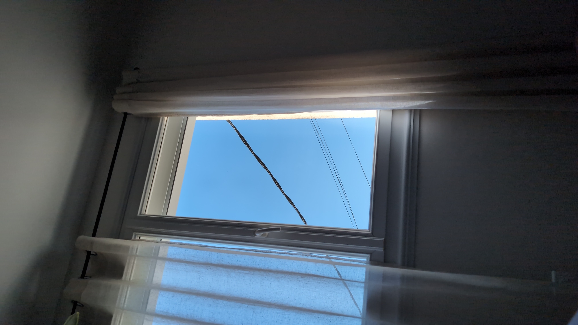 A view through a window in Cholet, France, reveals a bright blue sky with a few thin, dark wires crossing it. The window is framed by sheer white curtains that are partially drawn, allowing sunlight to filter through. The room itself is dimly lit, with shadows cast on the wall beneath the window. The overall impression is one of a quiet, domestic scene with a glimpse of the outside world.