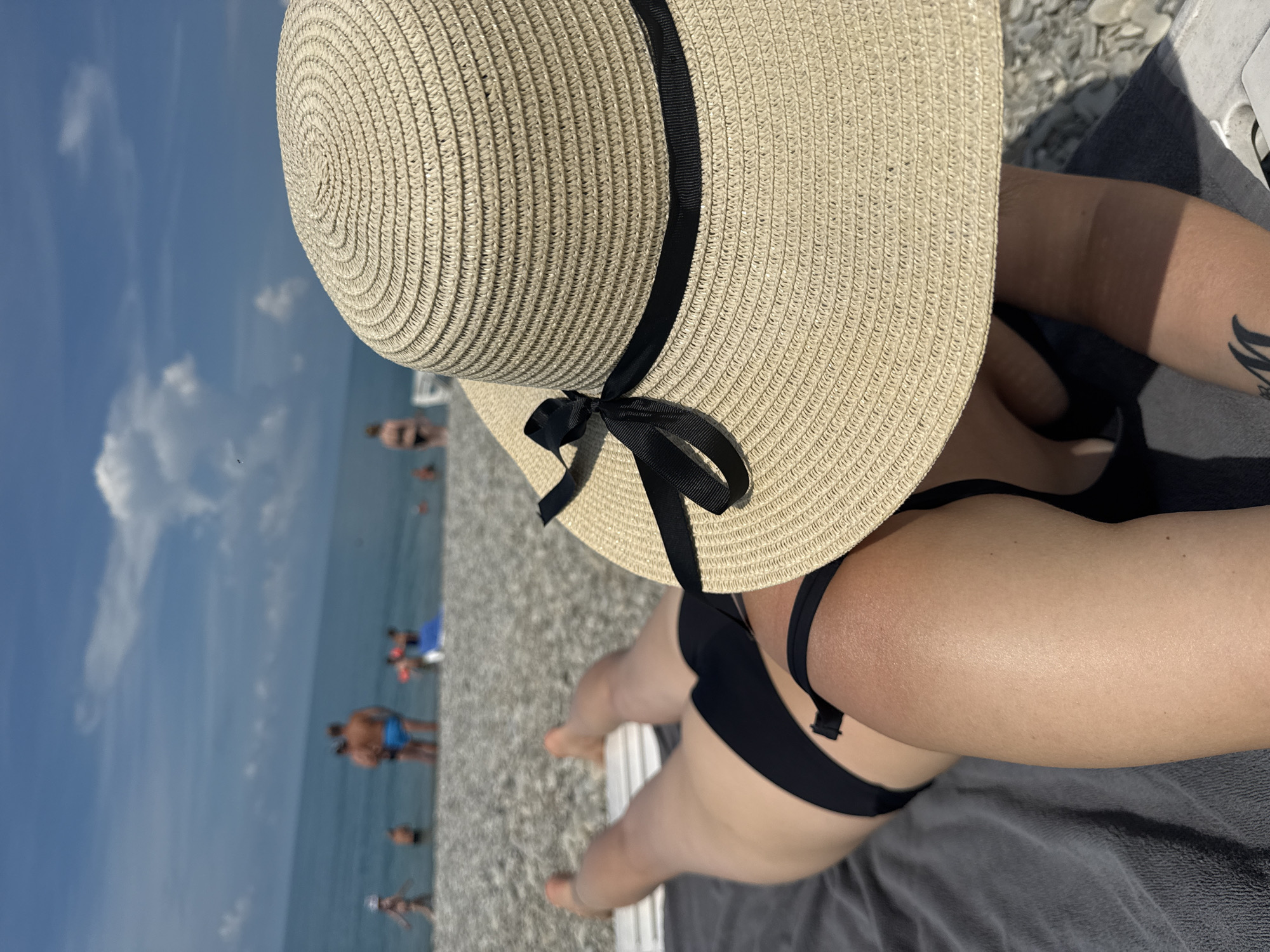 A person lies on a lounge chair on a pebble beach, wearing a black bikini and a straw hat with a black ribbon. The person's back and legs are visible, with a tattoo on their arm. In the background, the sea is calm under a blue sky with scattered clouds. Several people are visible in the water and on the beach. The scene is located on the coast of the Black Sea in Nebug, Russia, a resort village known for its pebble beaches and clear waters.