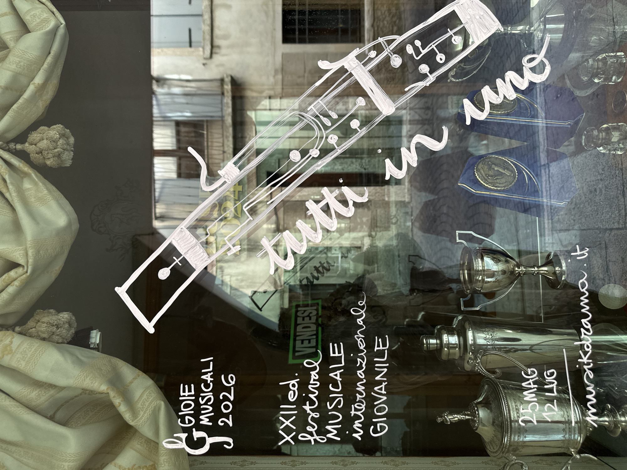 The image is a photograph taken at street level, looking through a shop window in Maser, Italy. White chalk art is drawn on the glass, depicting a musical instrument resembling a bassoon, along with text. To the left of the instrument drawing, text reads "GIOIE MUSICALI 2026" with a musical G clef. Below this, it says "XXII ed. festival MUSICALE internazionale GIOVANILE". A green sign in the background reads "VENDESI" (for sale).
Further down, on the left side of the window, a silver trophy is visible, with text written on it in chalk: "25MAG 12 LUG". To the right of this trophy is a silver shaker and another silver trophy. Behind these objects, two blue awards with gold medallions are displayed. In the bottom right corner, the text "musicheilrama.it" is written on the glass.
The shop window reflects the street outside, showing a building with balconies and windows, suggesting an urban or town setting. The overall lighting indicates it is daytime, with some natural light filtering through the window. The chalk art suggests the shop may be related to music or events.