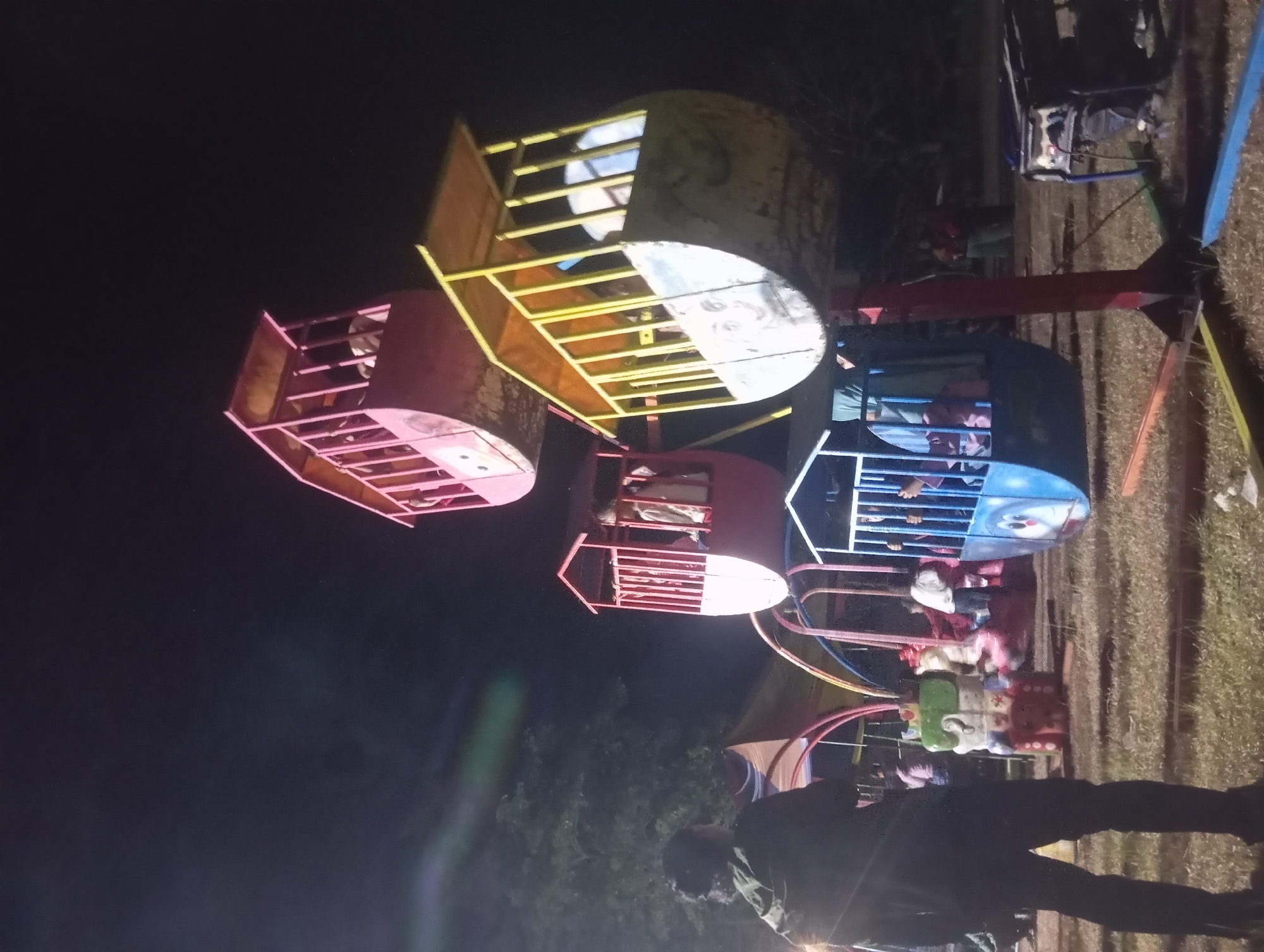 An outdoor amusement park scene at night in Dente Teladas, Indonesia, features a small Ferris wheel-like ride with brightly colored gondolas. The Ferris wheel structure is visible, with a person standing in the foreground on the left, facing away from the camera and towards the ride. Several gondolas are attached to the rotating mechanism. One gondola at the top is pink and appears empty. Below it, another gondola is yellow and shows a drawn-on face. A red gondola is positioned lower, with figures visible inside. At the bottom, a blue gondola also has figures. The ground is covered in dry grass, and a generator is visible on the right side of the frame. The background includes trees and a dark sky, illuminated by the lights of the amusement park.