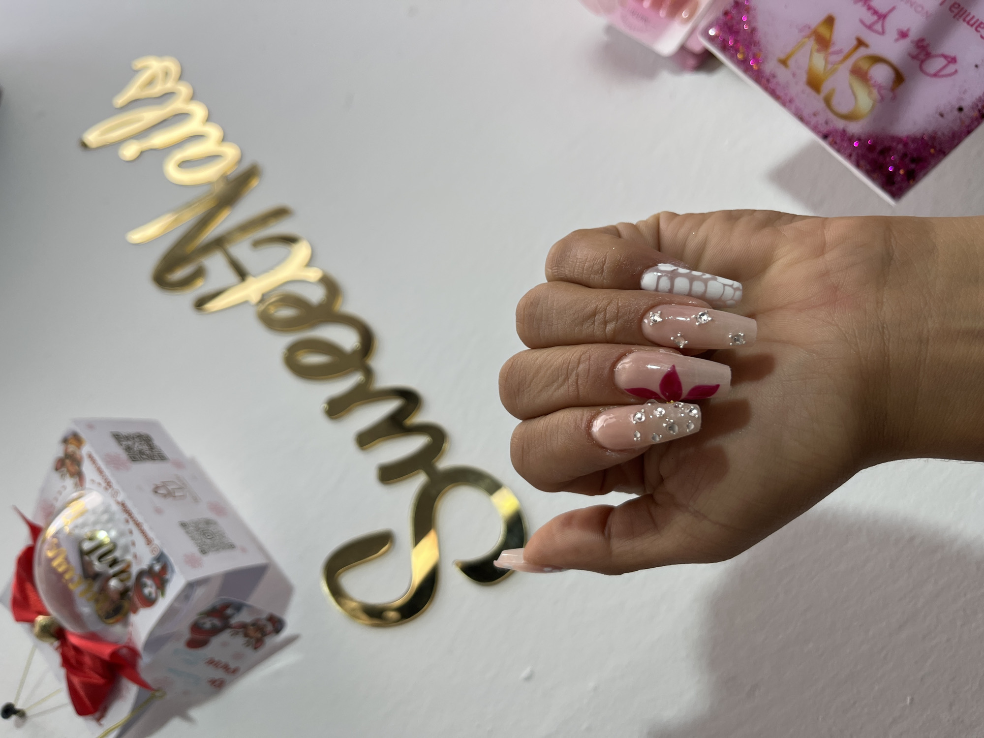 The image shows a hand with decorated fingernails. The nails are long and rectangular, with a nude base color. One nail is decorated with a white geometric design. Another nail has a pink flower design. The other nails have small crystals. The hand is positioned near a gold-colored "Queen Nails" sign, a box with a Christmas tree ornament inside, and a pink card with gold lettering. The scene is likely indoors. The location is Unknown, Unknown.