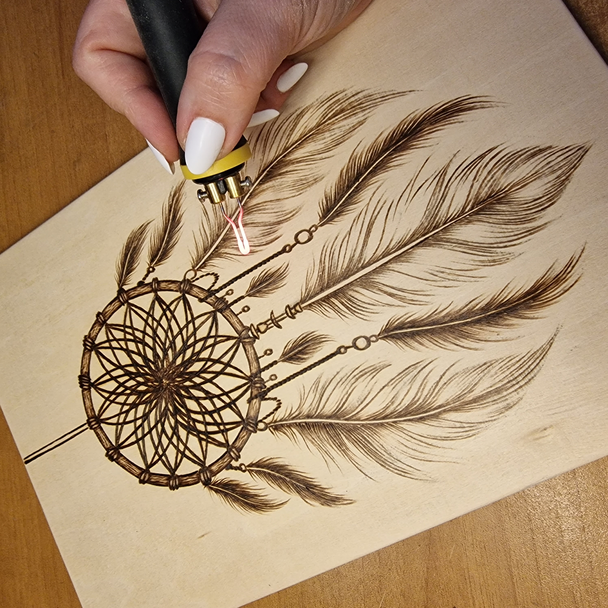 The image shows a close-up of a person's hand with white fingernails holding a wood-burning tool that is actively burning into a light-colored wooden surface. The tool's tip is glowing red and is being used to create a detailed drawing of a dreamcatcher with feathers. The wood-burning is in progress, with the intricate details of the dreamcatcher's web and the individual barbs of the feathers being etched into the wood. The background consists of a wooden table. There is no information in the image that indicates a location in Pleven, Bulgaria.