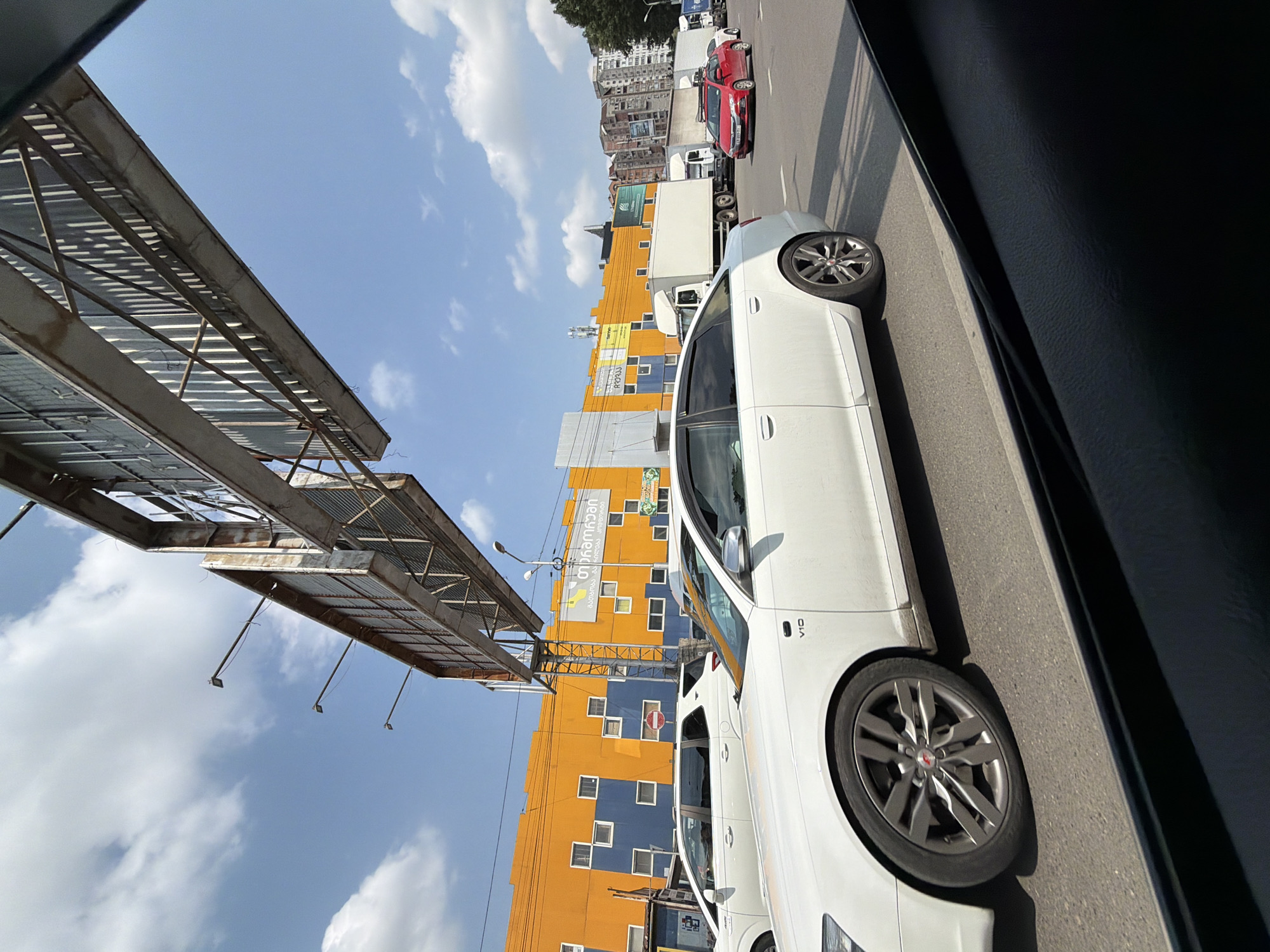 The image captures a street scene in Tbilisi, Georgia, under a clear sky with scattered clouds. In the foreground, the black interior of a car's window frame is visible. The main subject is a white Audi sedan, seen from the side, driving on a paved road. The car has a "V10" emblem on its side. To the left of the Audi, a white SUV is also visible.
In the background, an orange and blue building with a distinctive pattern of small square windows dominates the scene. A large, weathered billboard structure is suspended above the road, partially obscuring the sky. Further down the road, a line of white trucks and a red sedan are driving. Buildings of various heights and architectural styles, some appearing to be apartment blocks, are visible in the distance. The overall impression is of a typical urban environment with traffic and commercial buildings.