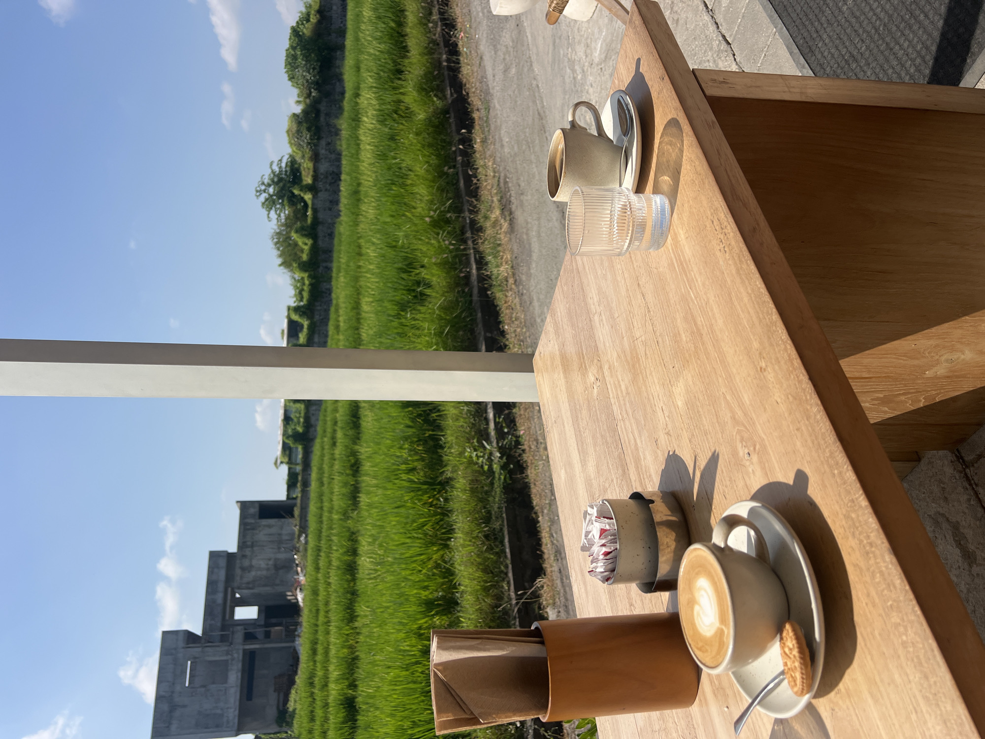 A wooden table is set with two cups of coffee, a glass of water, and a biscuit. In the background, a lush green rice paddy stretches towards a partially constructed concrete building. The sky is a clear blue with a few scattered clouds. The scene appears to be a cafe in Mengwi, Indonesia, offering a tranquil setting with views of the natural landscape and local development.