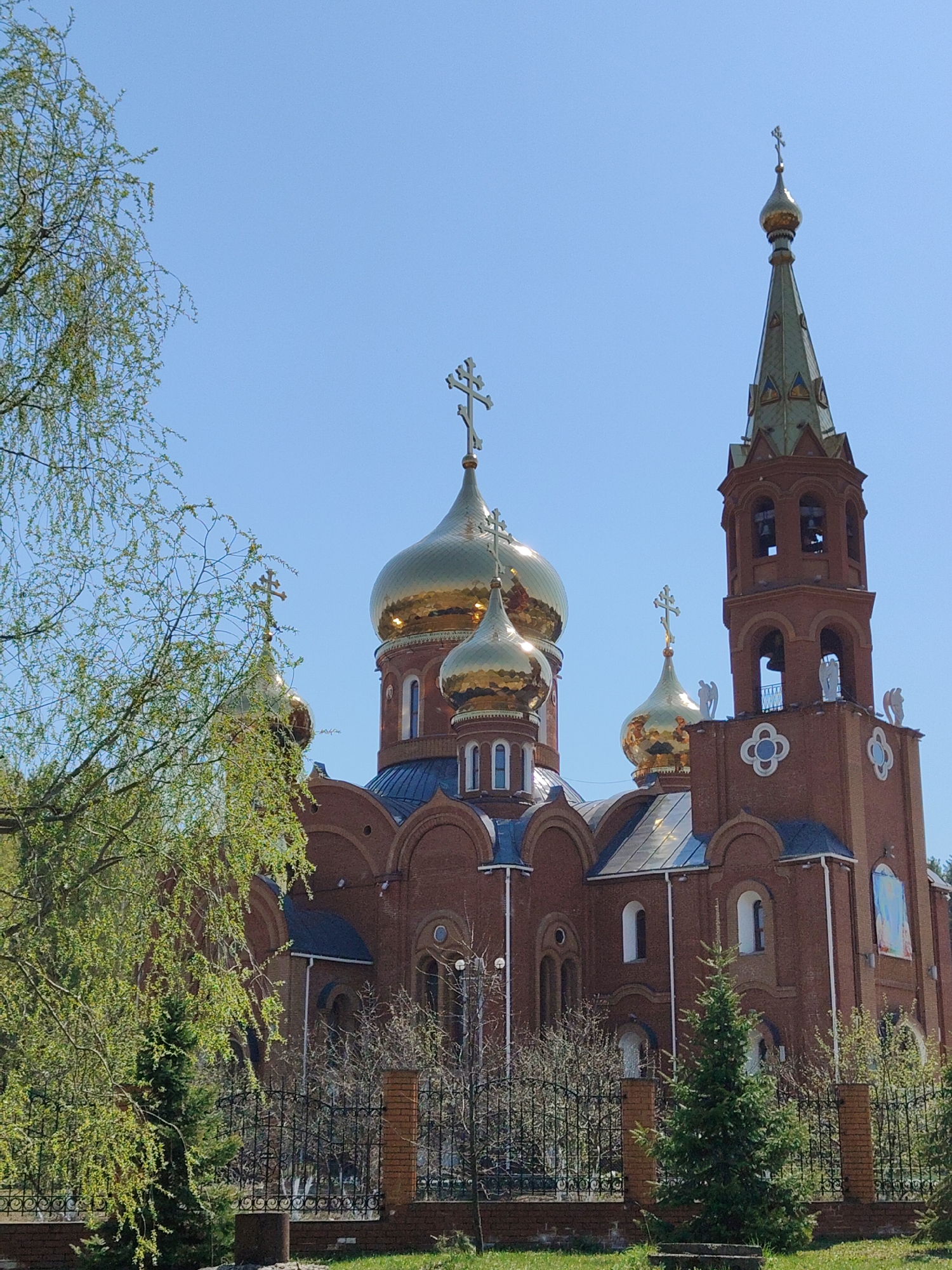 The image shows the Cathedral of All Saints in the city of Unknown, Unknown. The brick cathedral has multiple golden domes topped with crosses and a bell tower with a green spire. The cathedral is surrounded by a black metal fence and greenery. A tree with green leaves is visible on the left side of the image, and a small evergreen tree is in front of the fence. The sky is clear and blue.