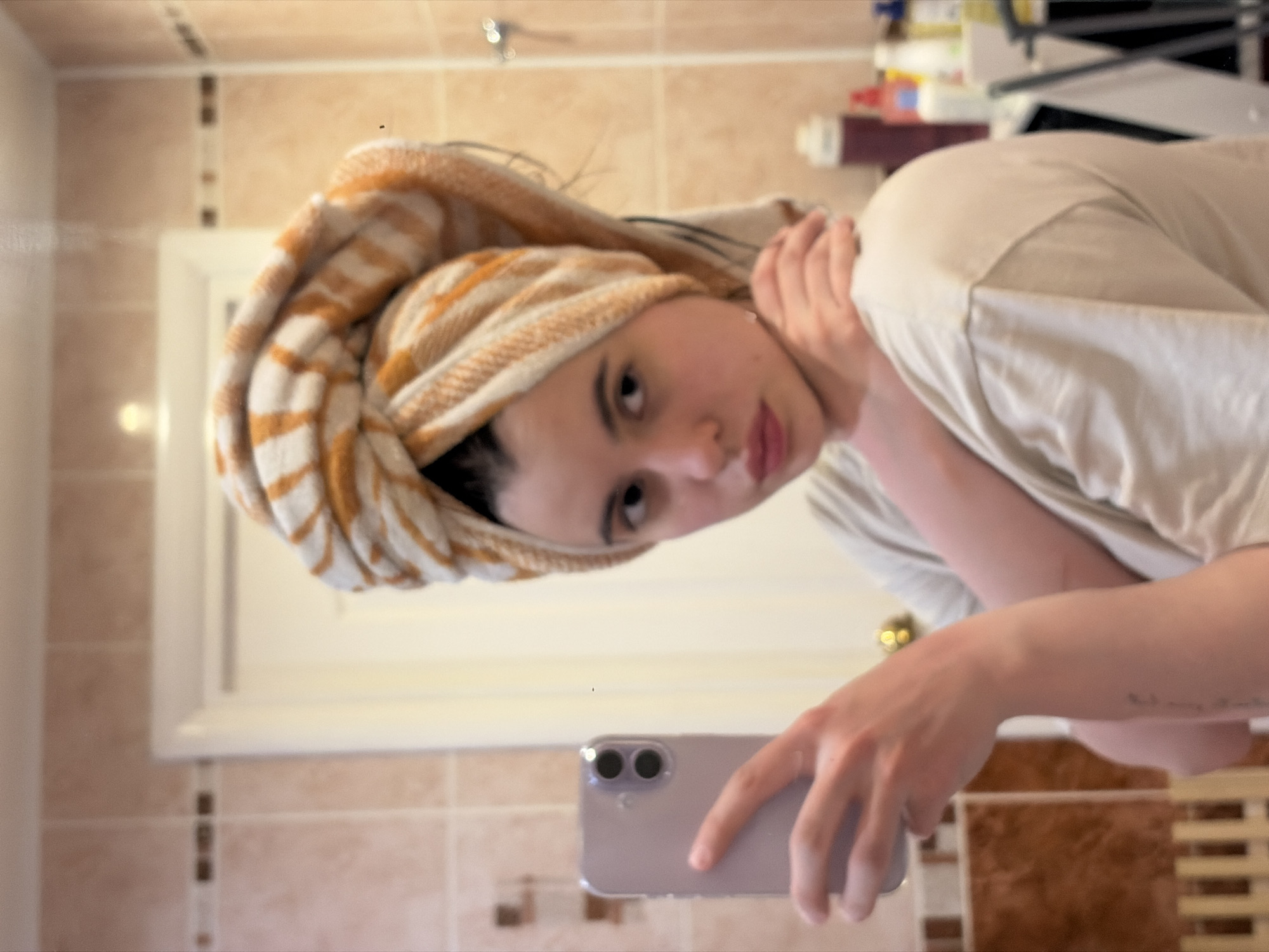 A person with a towel wrapped around their head is taking a selfie in a bathroom. The person is wearing a light-colored t-shirt and holding a mobile phone with dual cameras. The bathroom has light brown tiled walls with decorative inserts and a white door. The city of Chaikovsky, Russia is not discernible from this image.
