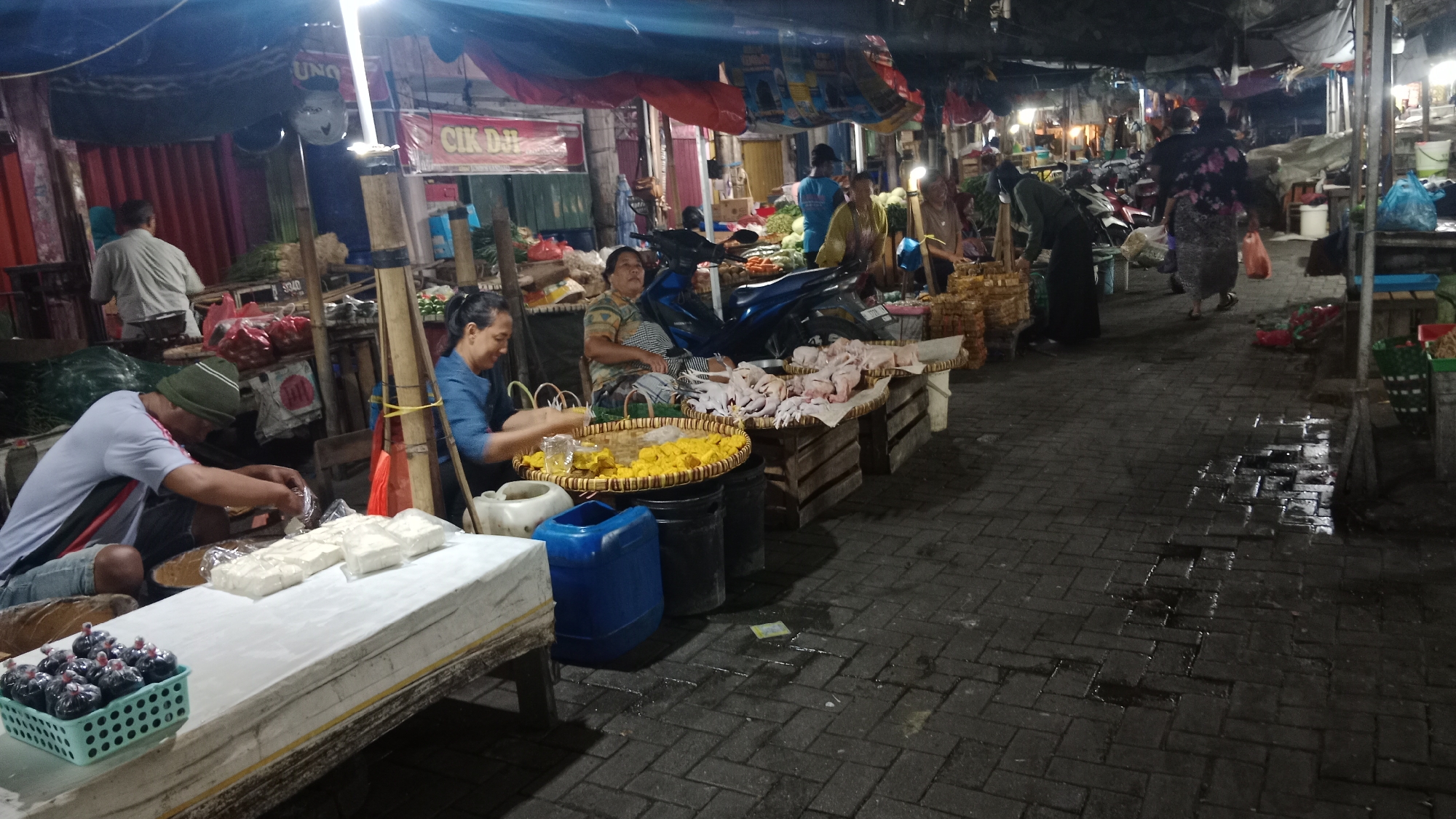 This is a medium shot of a bustling outdoor market in Semarang, Indonesia, at dusk. The foreground features a stall with white plastic-wrapped blocks of food, likely tofu, and a basket of small, dark-colored, round items, possibly preserved fruits or spices. A man wearing a green beanie and a grey shirt is seen from the side, actively packaging the white blocks of food. To his right, a woman in a blue shirt is sitting at a stall, her hands occupied with a yellow, diced food item spread on a woven mat. Behind her, another person in a striped shirt is seated, looking towards the foreground.
The market stalls are constructed from wood and bamboo, draped with tarps and awnings of various colors, including blue and red. Various goods are displayed, such as raw chicken parts on woven trays and bunches of fresh produce. A blue plastic container and black buckets are visible near the food stalls. The ground is paved with dark bricks, wet in patches, suggesting recent rain or cleaning. Several motorbikes are parked amongst the stalls, indicating a common mode of transport for vendors and customers. In the background, more stalls and people are dimly lit, hinting at the market's extensive size. The overall atmosphere is that of a lively, local marketplace engaged in daily commerce.