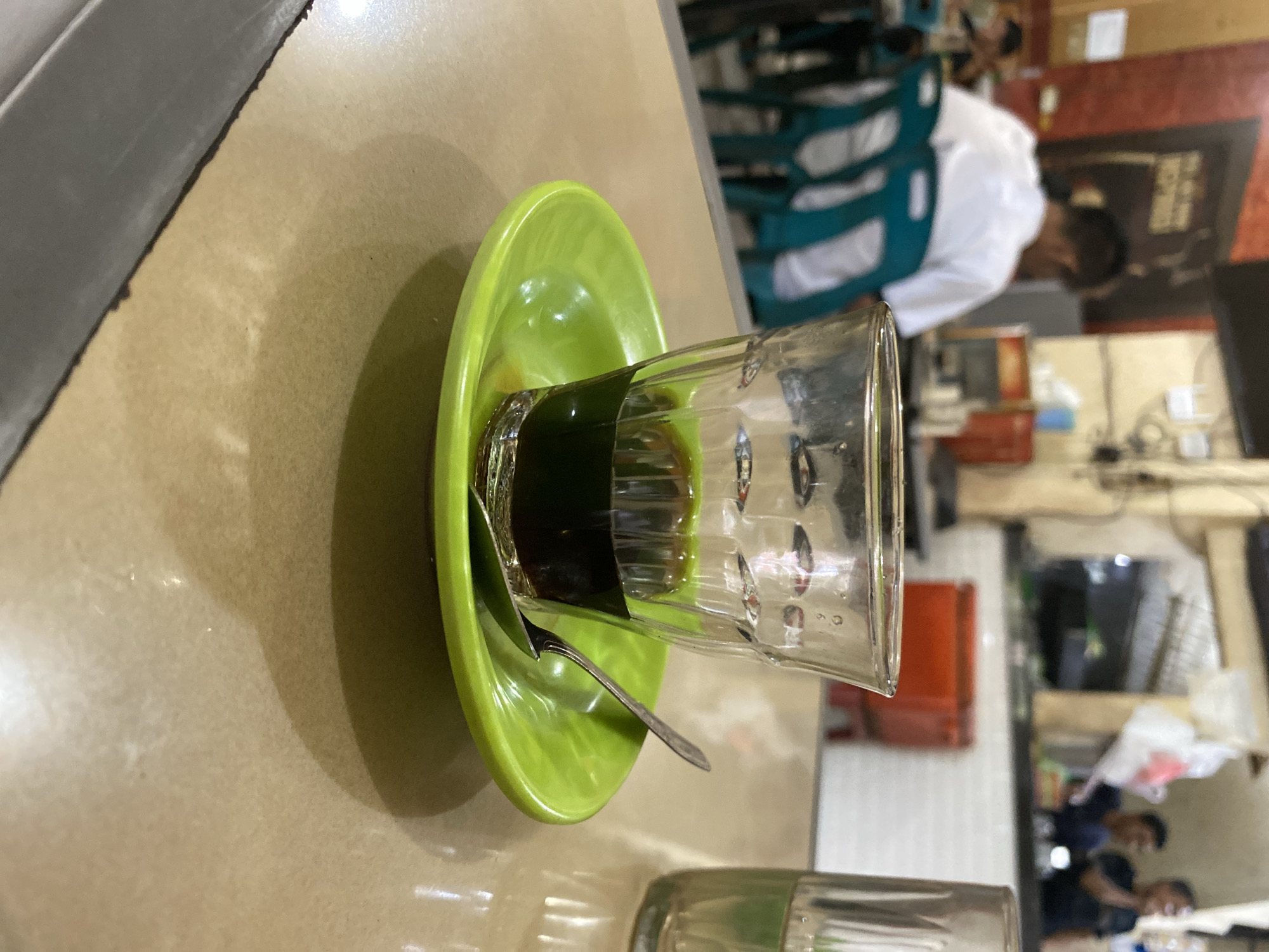 The image depicts a close-up view of a small glass of dark liquid, possibly coffee or tea, resting on a vibrant green saucer. A silver spoon is placed on the saucer, partially submerged in the liquid. The glass is made of clear, textured glass and has a slight ripple effect. The saucer is a bright, almost neon green plastic or melamine.
The background is blurred, suggesting a casual indoor setting, likely a cafe or a restaurant. In the distance, several people are visible. One person in the middle ground wears a white shirt and is seated, facing away from the camera. To the right, two men are visible behind a counter. One appears to be speaking while the other looks on. The overall ambiance suggests a lively but relaxed atmosphere.
The setting is identified as Lhokseumawe, Indonesia, implying a local eatery or coffee shop within that city. The lighting is warm, with some reflections visible on the tabletop, indicating an indoor environment with overhead or artificial lighting. The image is taken from a slightly elevated angle, focusing on the drink and saucer in the foreground.