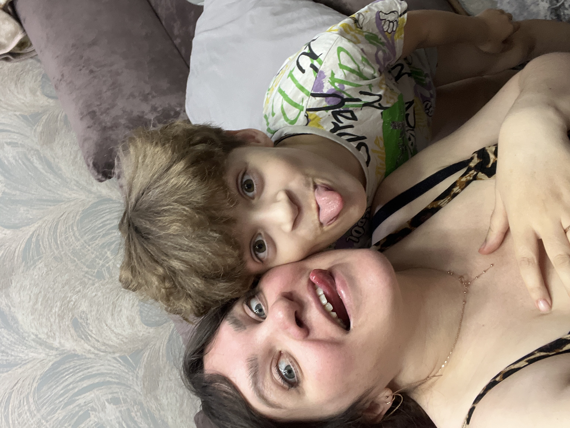 A woman and a child are lying down on a surface with a patterned design. The woman has dark hair and is wearing a top with animal print straps and a gold necklace. She is looking upwards with her mouth open and tongue out. The child, with curly brown hair, is lying above her, also sticking out his tongue. The setting appears to be indoors, possibly on a bed or couch. No city of Noviy, Russia is evident in the image.