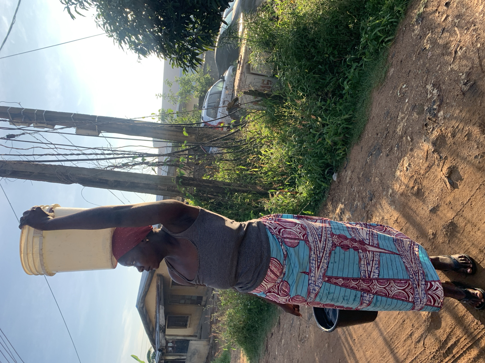 A woman is shown from the knees up, carrying a large yellow bucket of water on her head. She is wearing a grey tank top and a blue and red patterned skirt. She is also carrying a black bucket in her left hand. She is walking on a dirt road in a rural area. In the background, there is a house and some trees. The sky is clear and blue. The image appears to have been taken in the city of Rafin Par, Nigeria.