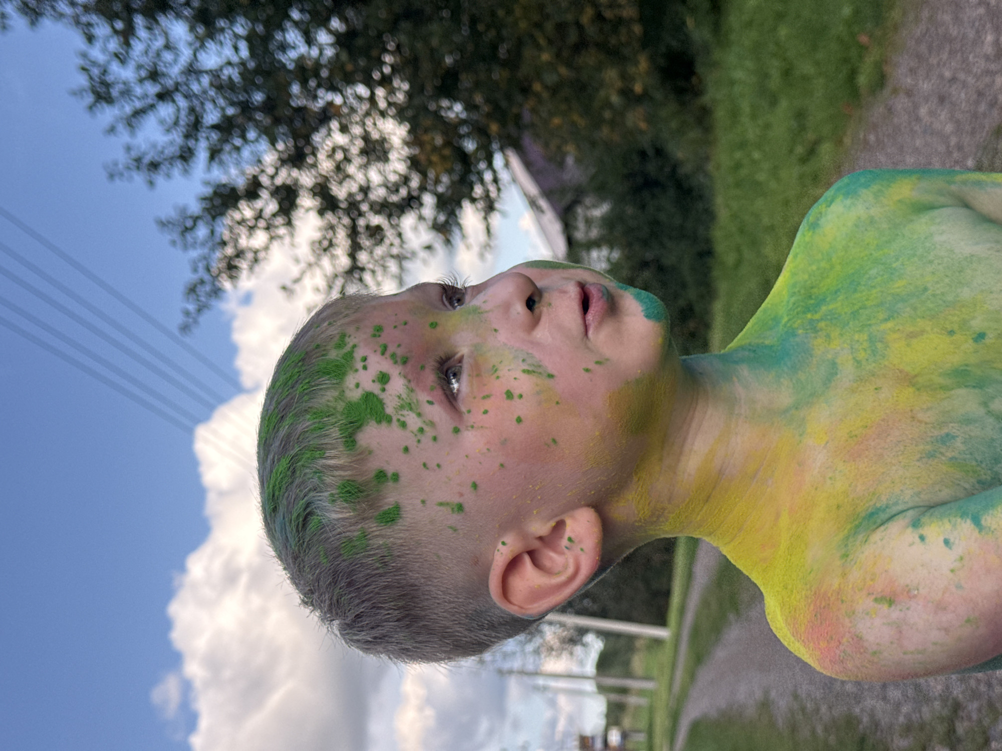A young boy, with his face and upper body covered in green, yellow, and some pink powder, looks to his right. He has short, light brown hair with green powder splotches. His mouth is slightly open, and his eyes are looking upwards and to the right. The background shows a dirt path leading away into a grassy area, with trees and a building visible in the distance under a cloudy sky. Overhead power lines are also present. The setting appears to be a rural outdoor location, possibly Nizhnyaya Chermoda, Russia, given the provided context. The boy is shirtless, and the powder is applied unevenly, creating a playful and colorful effect.