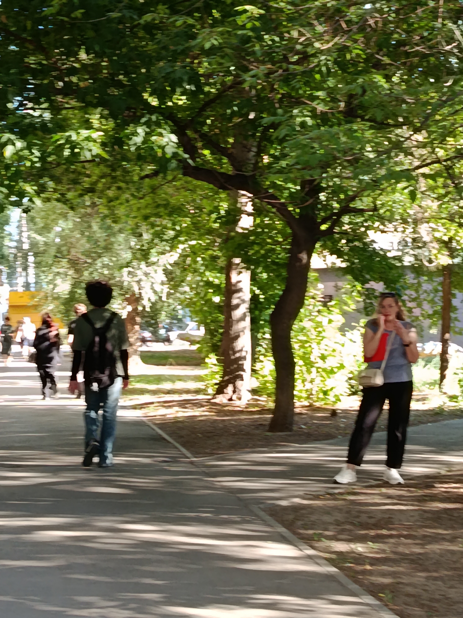 On a sunny day, several people walk along a paved pathway lined with trees. In the foreground, a person with dark hair wearing a green shirt and backpack walks away from the camera. To the right, a woman with blonde hair, wearing a blue shirt and black pants, stands with a red object in her hands and a small purse over her shoulder. In the background, other individuals are visible walking. The scene appears to be a park or a tree-lined street in Saratov, Russia.