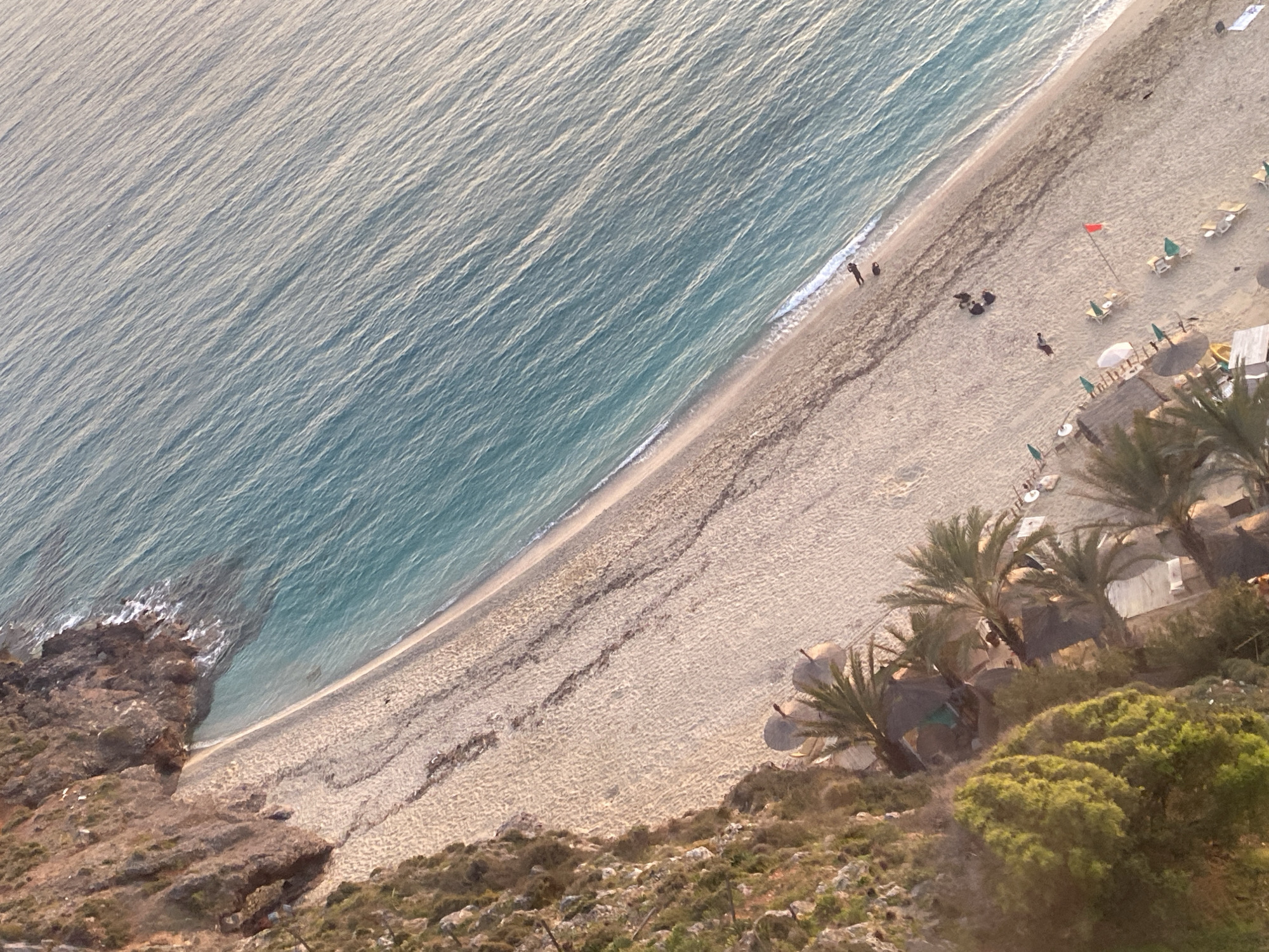 The image presents an aerial view of a beach in Alanya, Turkey. The scene includes the Mediterranean Sea, a sandy beach, coastal vegetation, and rocky terrain. The sea occupies the upper left portion of the frame, exhibiting a gradient of blue hues. The beach stretches along the coast, bordered by a strip of darker sand. On the beach, there are several sun loungers, beach umbrellas, and at least one red warning flag. A few individuals are visible on the beach, some walking along the water's edge. The lower portion of the image showcases the rocky terrain and sparse vegetation, indicative of the Alanya coastline. Palm trees are visible near beach umbrellas, likely providing shade.