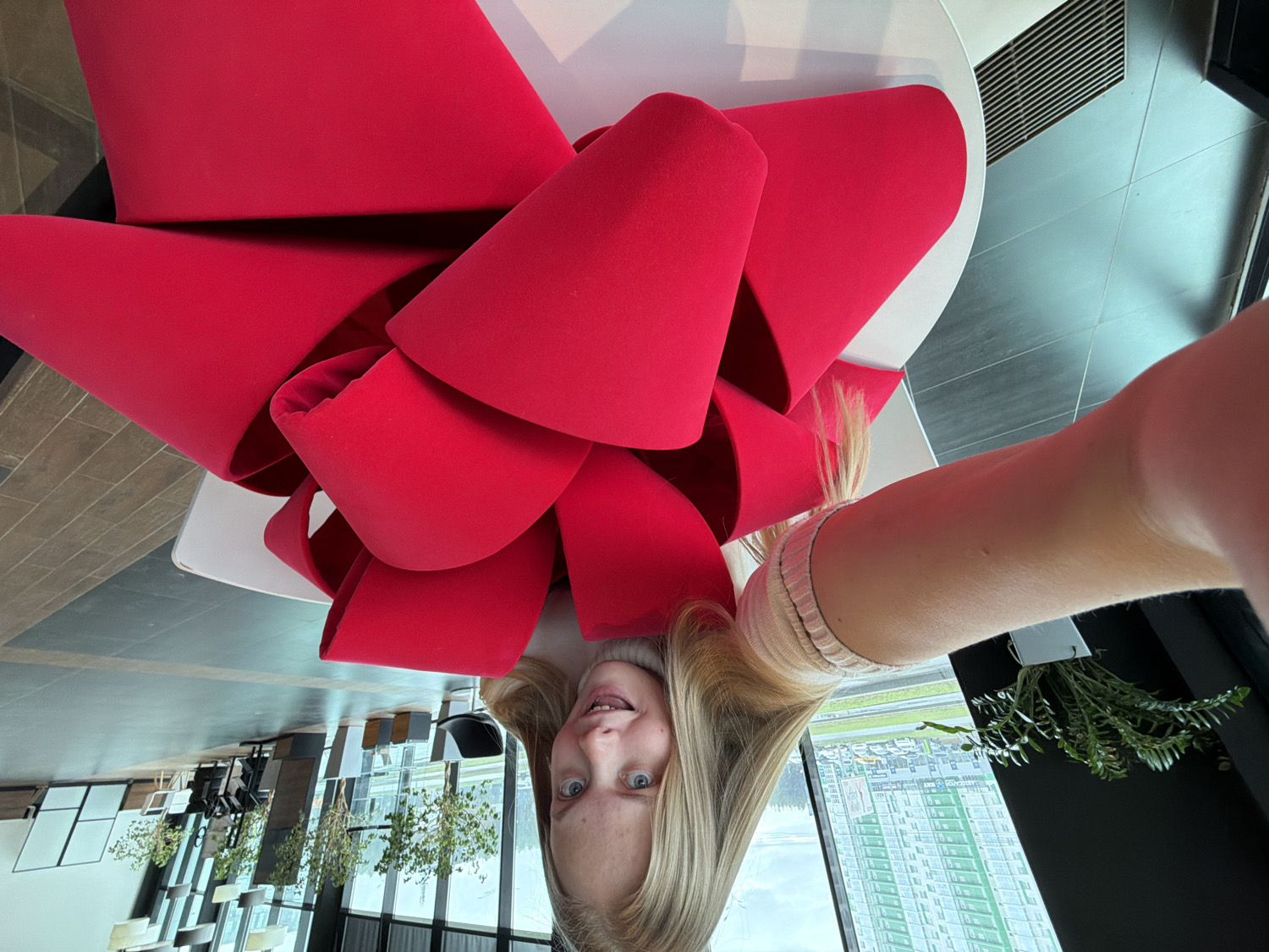 The media file presents an upside-down selfie of a young person in a modern interior. The top half of the image is dominated by a large, sculptural object in a vivid, matte red color, resembling a colossal fabric bow or an abstract piece of furniture. It appears to be made of a soft material like felt or velour and rests on a white, circular surface.
The bottom half of the image features the face of a young individual, likely female, with fair skin, light blue eyes, and long blonde hair, smiling subtly at the camera. Their right arm and hand are visible extending from the right side of the frame, indicating they are holding the device for the selfie. A light-colored, possibly silver-toned, bracelet is visible on their wrist.
The background behind the person, viewed as if reflected or seen through large windows, reveals a spacious, contemporary indoor setting. Elements such as modern tables, chairs, and several potted green plants are discernible. Beyond the windows, an urban landscape extends, featuring numerous tall buildings under what appears to be a bright, possibly overcast, daytime sky, consistent with a city environment like Izhevsk, Russia. The playful orientation of the selfie suggests a casual and perhaps artistic moment. No visible text is present in the image.
