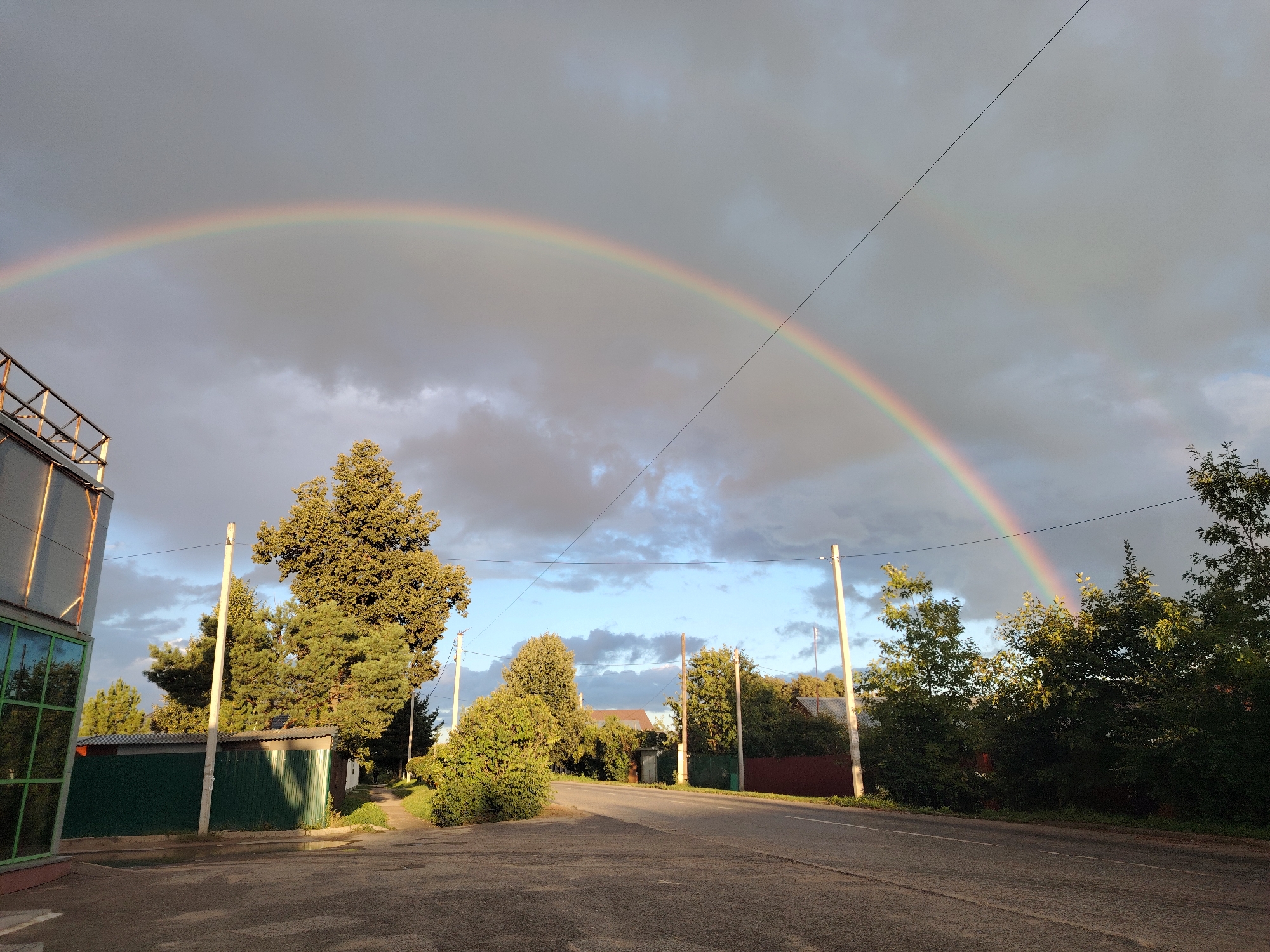 A wide shot captures a vibrant double rainbow arching across a cloudy, grey sky over a rural landscape. The left side of the image features a modern building with glass windows and a metal exterior, partially shaded by the overhead structure. A paved road curves through the scene, bordered by various trees and vegetation. Several utility poles with wires are scattered throughout, with one prominent pole on the left side and another to the right. In the foreground, the road appears to be made of asphalt, with some debris and puddles on its surface. The overall atmosphere suggests a post-rain scene, with the sun breaking through the clouds to create the stunning rainbow display. The environment appears to be a suburban or rural area, with hints of houses and fences visible among the trees in the background. No people or animals are present in the image.