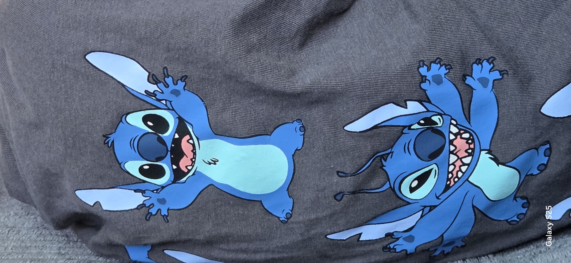 This is a close-up, eye-level shot of a dark grey t-shirt with two cartoon characters printed on it. The characters are blue, resembling Stitch from Disney's Lilo & Stitch. The top Stitch is positioned higher in the frame and appears to be jumping with its arms raised. The bottom Stitch is slightly lower and also has its arms raised, with a wide, toothy grin. The t-shirt fabric has a visible ribbed texture. The image does not contain any elements that would indicate a specific location in Eeklo, Belgium.