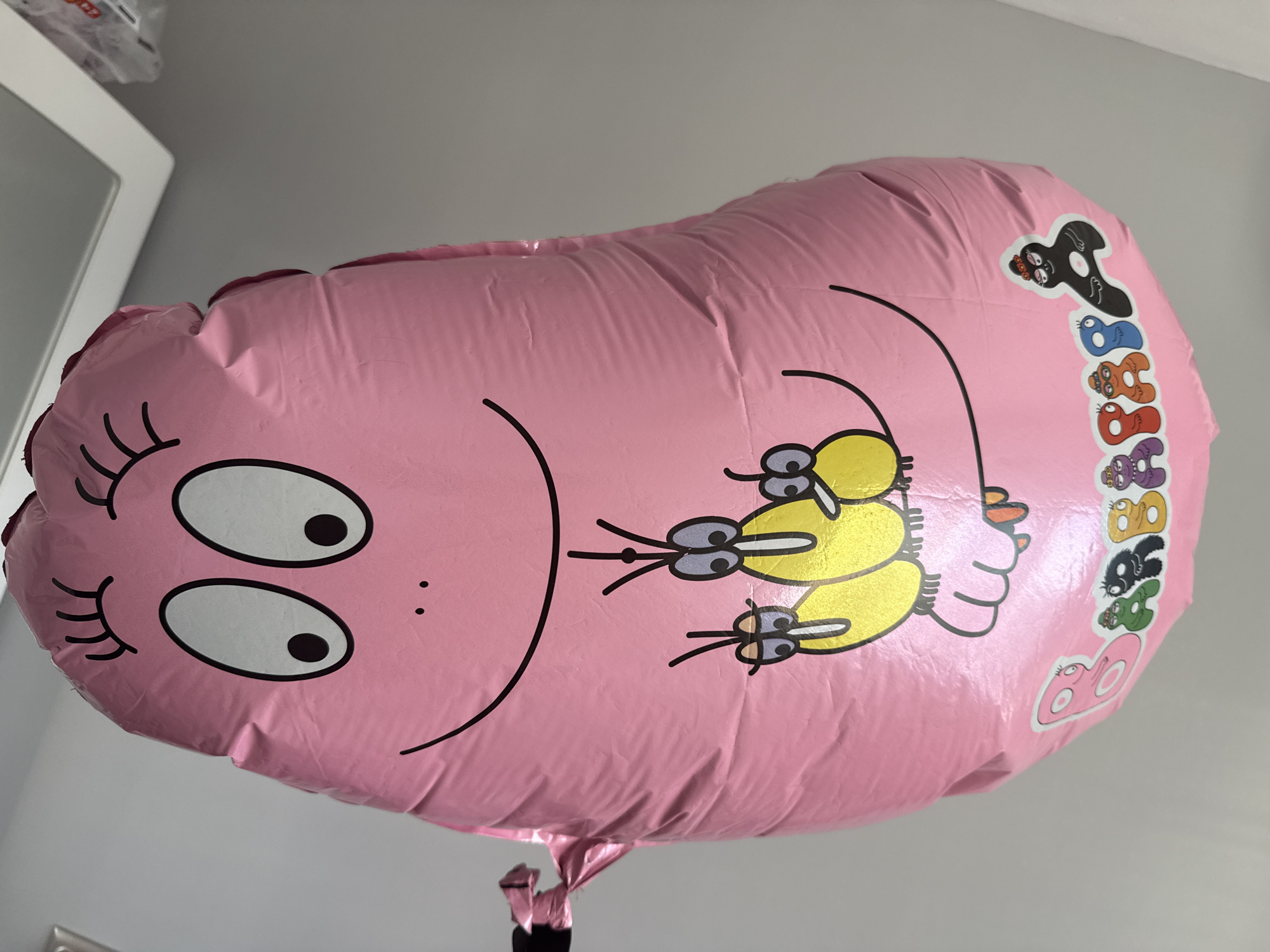 A pink, Mylar balloon in the shape of a character's head is suspended against a plain gray wall. The balloon features a cartoon face with closed, curved eyelashes on the left and two large, oval eyes with black pupils on the right. A simple smile is drawn beneath the eyes. Two yellow, insect-like creatures with antennae and large eyes are depicted on the right side of the balloon's face. Along the right edge of the balloon, a vertical sequence of colorful, cartoon-like characters is printed, beginning with a black character at the top and continuing with blue, orange, red, and purple characters below. The balloon is tied to a black string with a pink, reflective attachment at the bottom. The lighting is even, and the background is unremarkable, offering no contextual clues about its location or time of day. No people are visible in the image, and no actions are occurring. The scene is static and appears to be an indoor photograph. The balloon is the sole subject of interest. There are no details in the image that suggest a location in Burgas, Bulgaria.