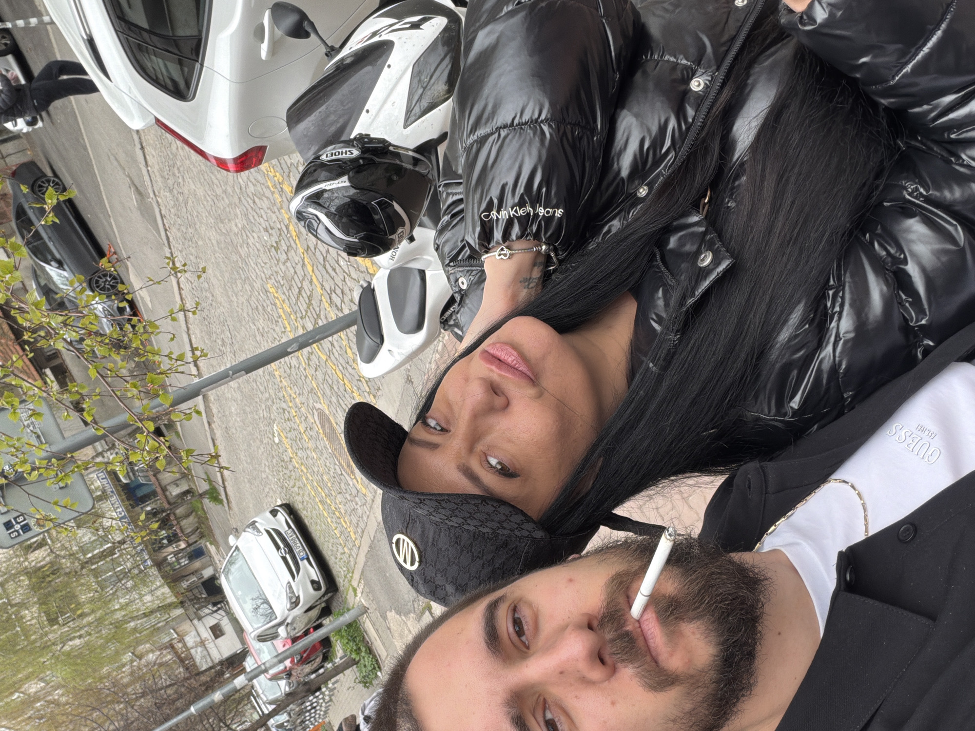 A street scene in Sofia, Bulgaria. Two people, a woman and a man, are posing for a photograph. The woman is wearing a shiny black puffer jacket with a "Calvin Klein Jeans" logo visible, and a black baseball cap with a white logo. She has long black hair. The man, positioned lower in the frame, has a beard and is wearing a white t-shirt with "GUESS" printed on it. He is also wearing a black blazer and smoking a cigarette. A white motorcycle with a white and black helmet is near the woman. In the background, there are cars parked along a cobbled street, and buildings can be seen in the distance.