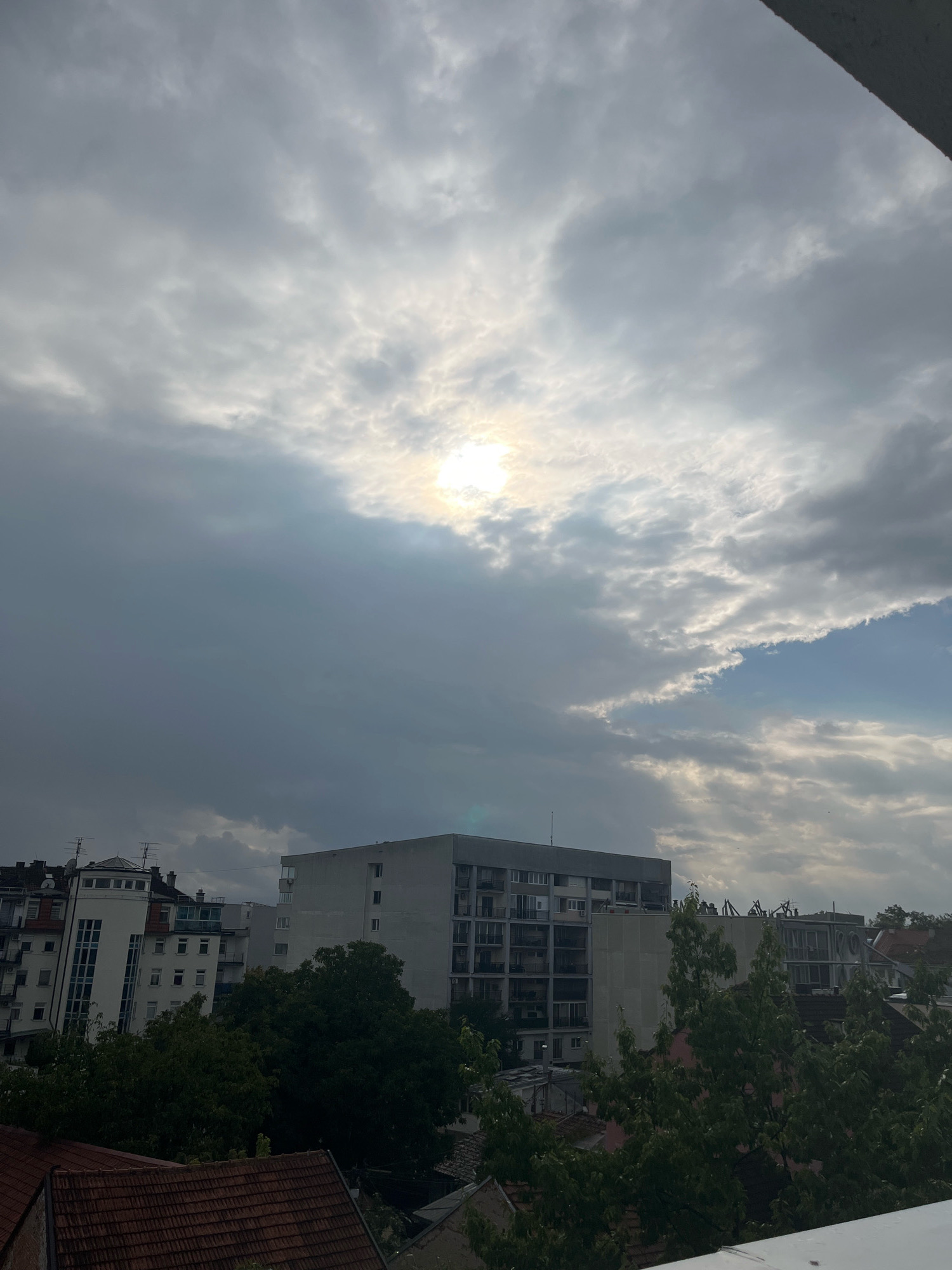 The image captures a cityscape in Kraljevo, Serbia, under a heavily overcast sky with the sun partially obscured by clouds. In the foreground, the tops of terracotta-tiled roofs are visible. Behind them, a dense canopy of dark green trees obscures the lower levels of buildings.
The midground is dominated by several multi-story buildings. A large, rectangular grey building with multiple balconies occupies a significant portion of the frame. To its left, a more modern white building with a curved facade and dark windows stands out. Further to the left and in the background, other residential buildings with visible windows and antennas are present. The sky is filled with dramatic clouds, ranging from dark grey to lighter, illuminated patches where the sun attempts to break through. The overall impression is a cloudy, possibly pre-rain, urban environment. There are no people or overt actions depicted in the scene.