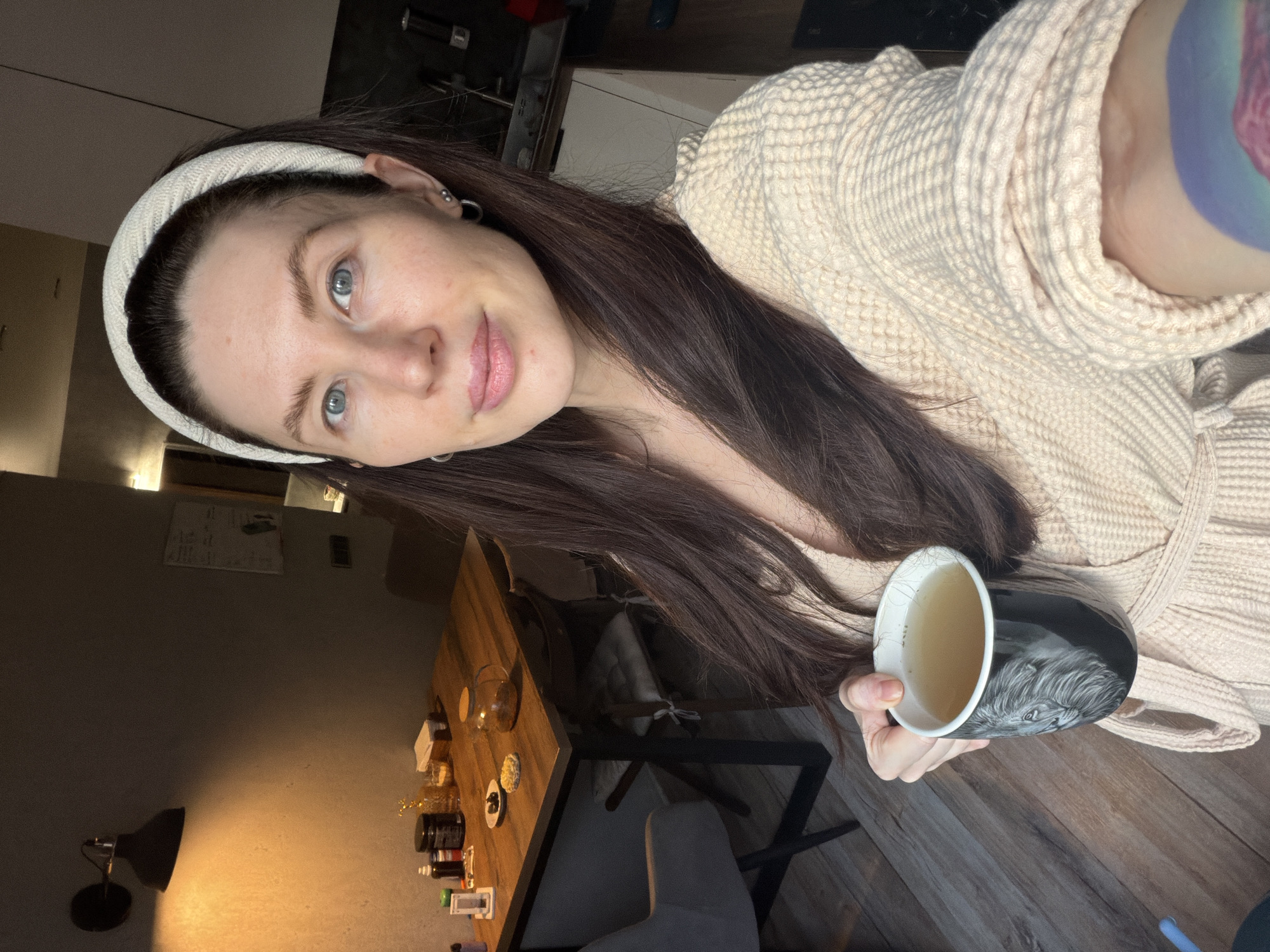 A person wearing a light beige bathrobe and a headband is holding a mug containing a light-colored liquid. The individual has fair skin, dark brown hair, and light blue eyes. The background features a wooden table with various small items on it, a chair, a lamp, and part of a wall. The setting appears to be indoors, possibly a residential space. The location is Perm, Russia.