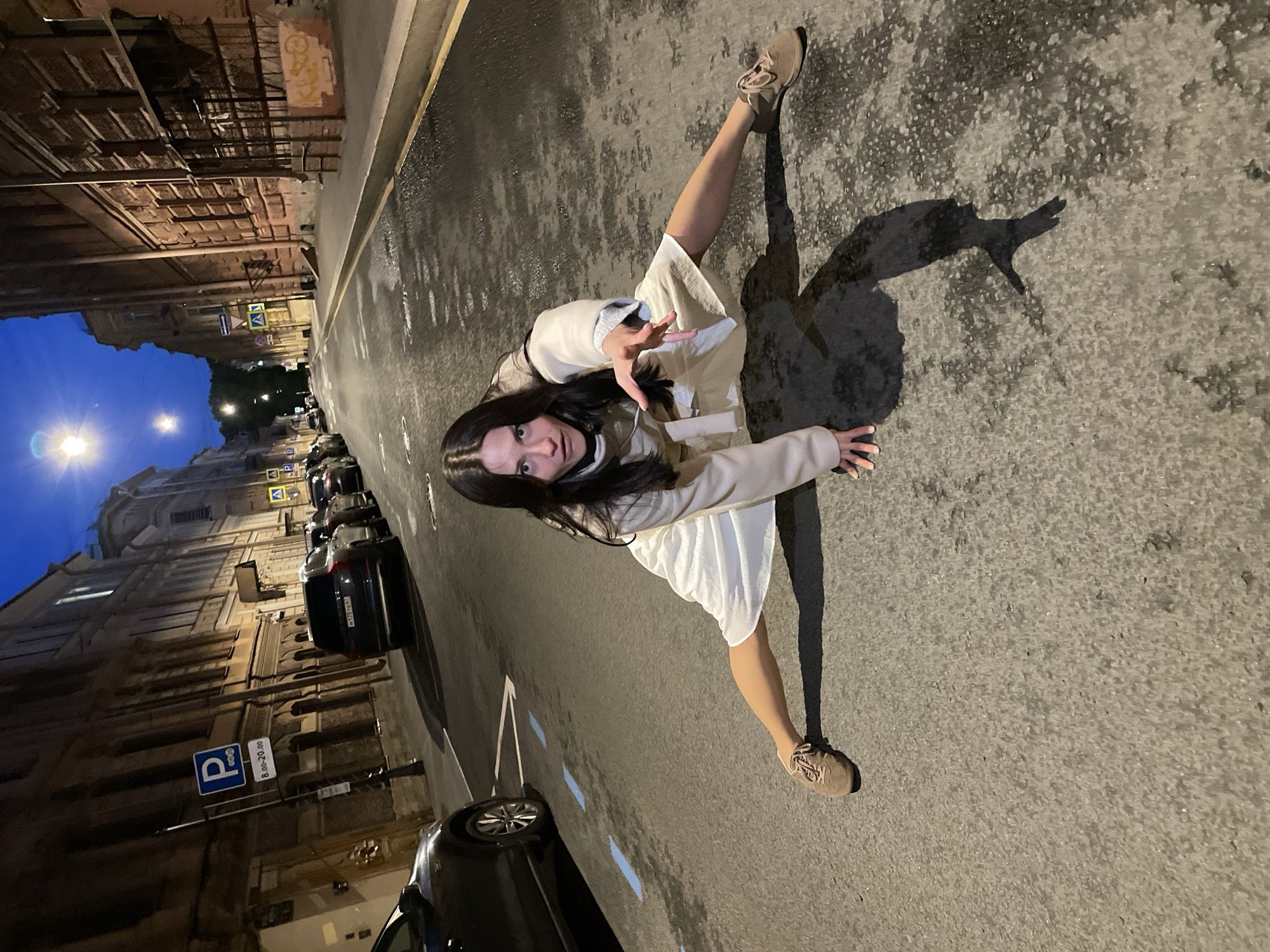 A young woman is on all fours in the middle of an asphalt street in Saint Petersburg, Russia. She is wearing a light-colored trench coat, a white dress, and brown sneakers. Her legs are spread wide in a straddle position, and her arms are outstretched. The street is wet, and streetlights cast long shadows. Buildings with arched windows and classical architecture line the street. Several cars are parked along the side of the road. A blue and white parking sign is visible on the left side of the image. The sky is dark blue, indicating it is nighttime.