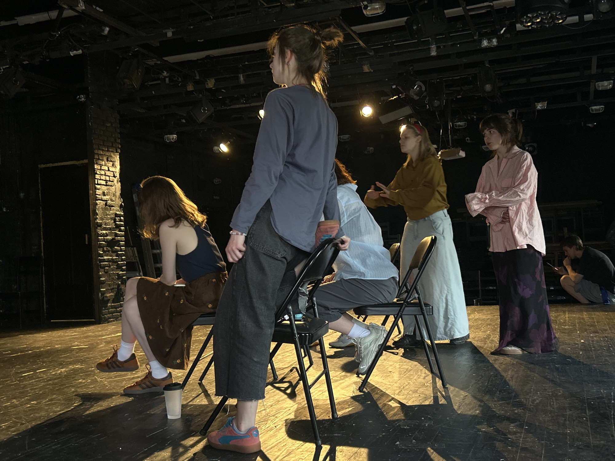 The image shows a group of people, mostly women, in a room resembling a theater or performance space. The location appears to be indoors with a dark, somewhat industrial aesthetic, including exposed pipes and lighting fixtures on the ceiling.
The people are positioned in a line, mostly seated on black metal folding chairs. From left to right:
1. A woman with long brown hair, wearing a navy tank top and a brown floral skirt with brown Adidas sneakers, is seated on a chair.
2. A woman is standing with her back to the camera, wearing a long-sleeved grey shirt and dark denim pants.
3. A woman wearing a light blue shirt and grey pants is seated on a chair.
4. A woman wearing a tan long-sleeved shirt and a white maxi skirt is standing. Her hair is styled with a red headband.
5. A woman wearing a pink striped shirt and a dark floral maxi skirt is standing.
To the right of the group, in the background, a person in a dark shirt and shorts is crouched on the floor.
The floor is dark and appears to be made of wood, with visible shadows and scuff marks. A plastic cup sits on the floor next to the woman on the left. The room is dimly lit by several overhead lights, which cast strong shadows.