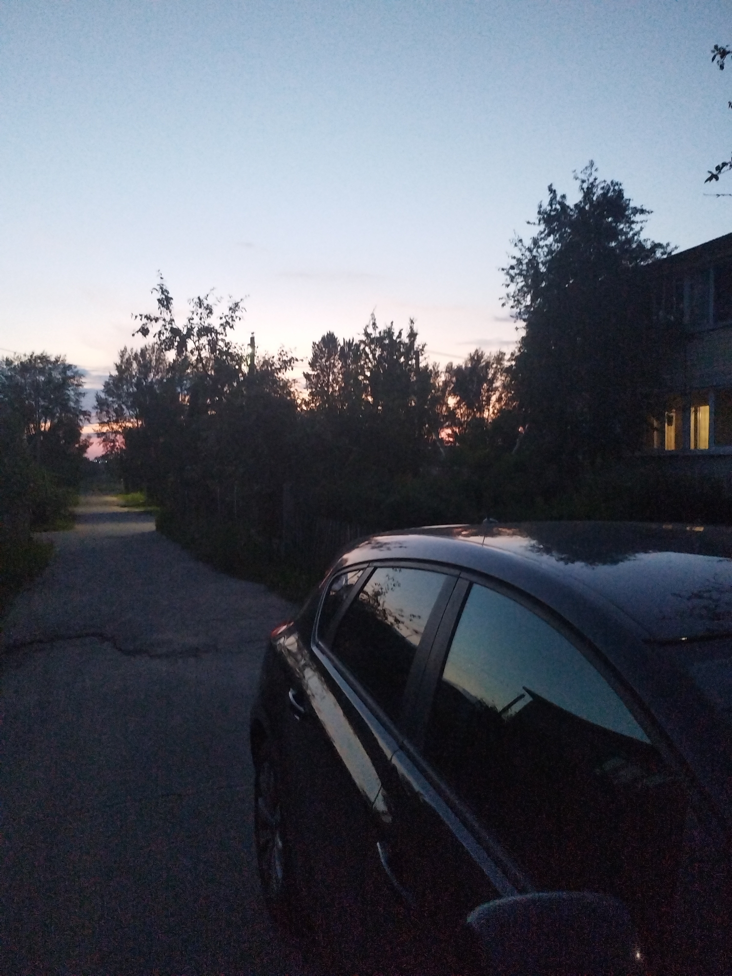 A dark blue car is parked on the side of a cracked asphalt road as dusk settles in. The sky above is a pale blue, transitioning to a faint pink near the horizon, indicating the end of daylight. Silhouetted trees line the road, some appearing dense with foliage, others more sparse. In the background, a building with lit windows is visible, suggesting it is inhabited. The overall atmosphere is quiet and serene, characteristic of an evening in a suburban or rural setting.