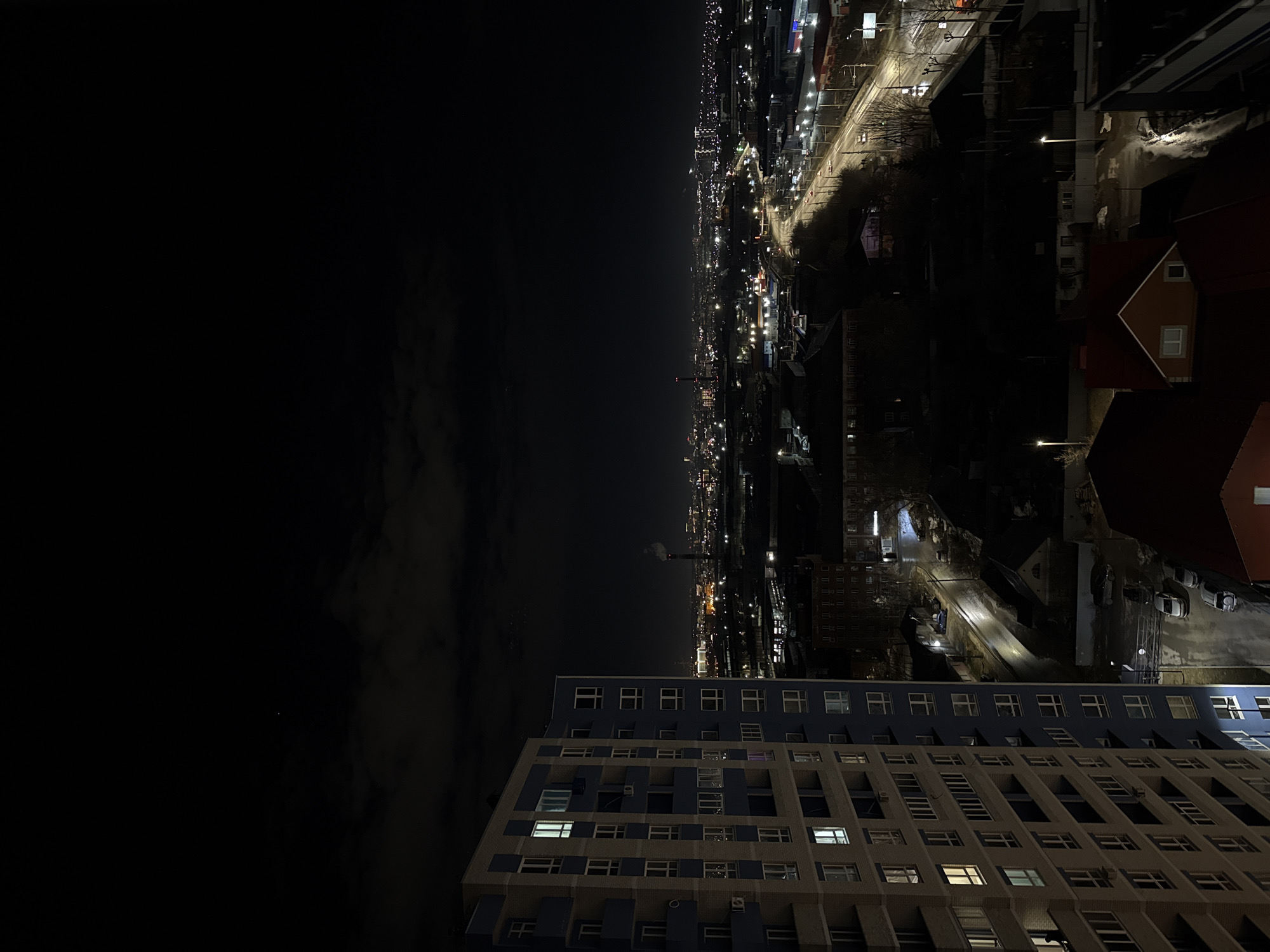 The image shows a nighttime view of Izhevsk, Russia. In the foreground is a multi-story building with a grid of windows, some lit and some dark. Beyond the building, the cityscape stretches into the distance, with streetlights illuminating roads and smaller buildings. The night sky is dark, with some clouds visible in the upper left portion of the image.